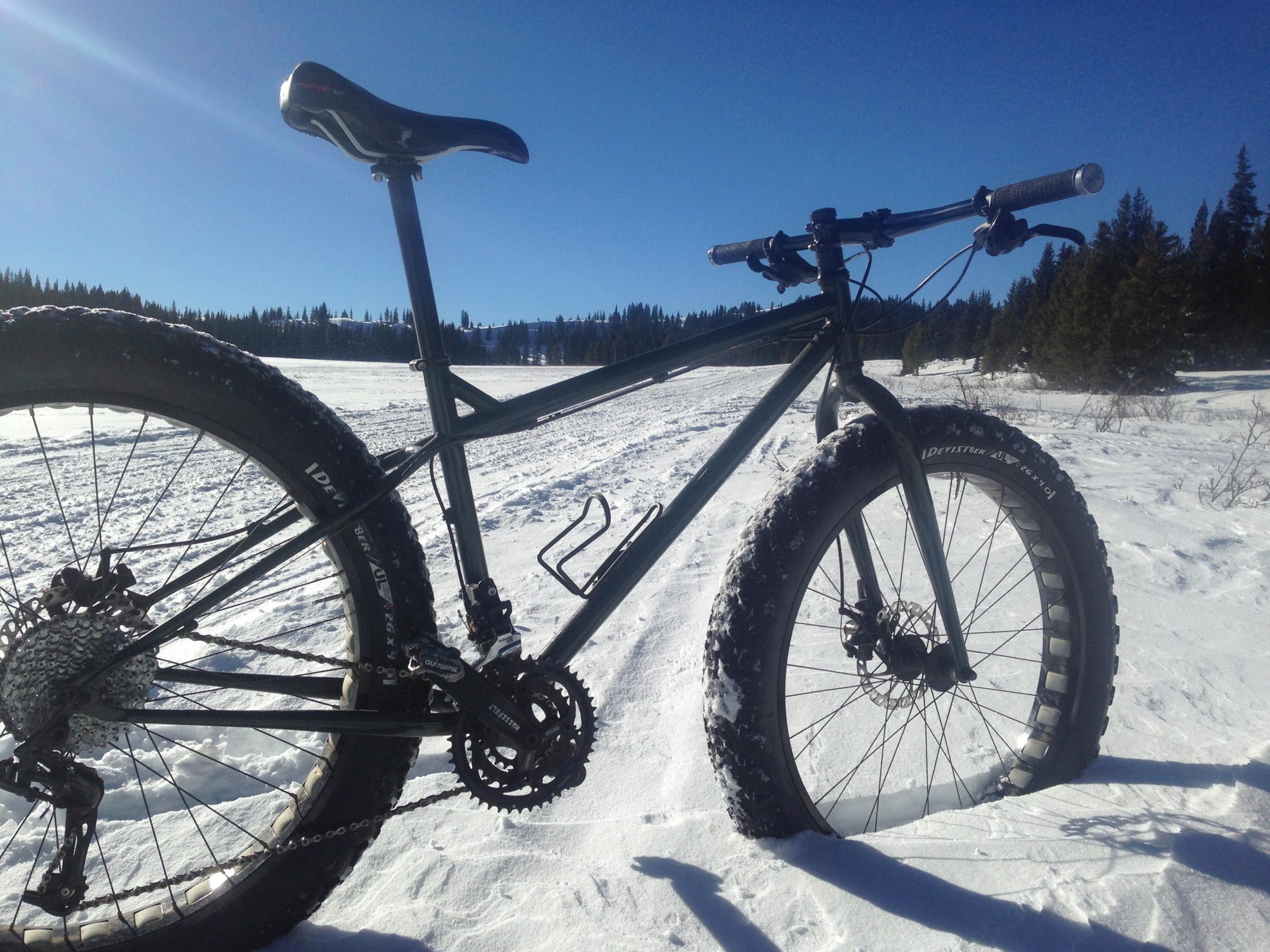 A fat tire bike parked on a snowy landscape, with a clear blue sky overhead. The bike features large, knobby tires designed for snow riding, a sleek frame, and prominent gears. In the background, snow-covered terrain and evergreen trees are visible, creating a serene winter scene. Marshall Pass Road / #200 / #203 / #243 mountain bike trail.