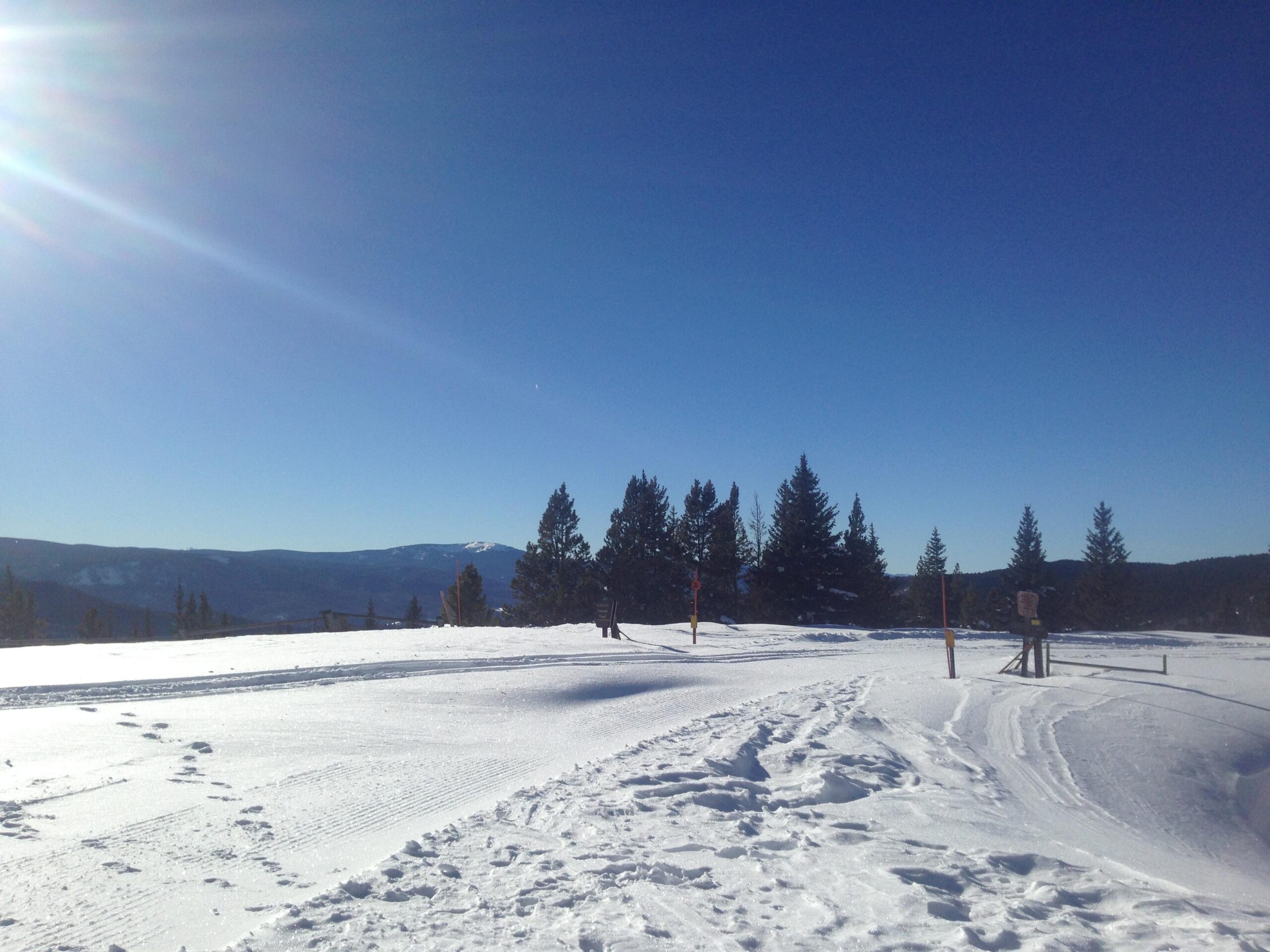 A snowy landscape under a clear blue sky, featuring a bright sun in the upper left corner. The scene includes tall evergreen trees in the background and a snowy foreground with visible tracks and trails. A mountain range is visible in the distance, completing the serene winter scenery. Marshall Pass Road / #200 / #203 / #243 mountain bike trail.
