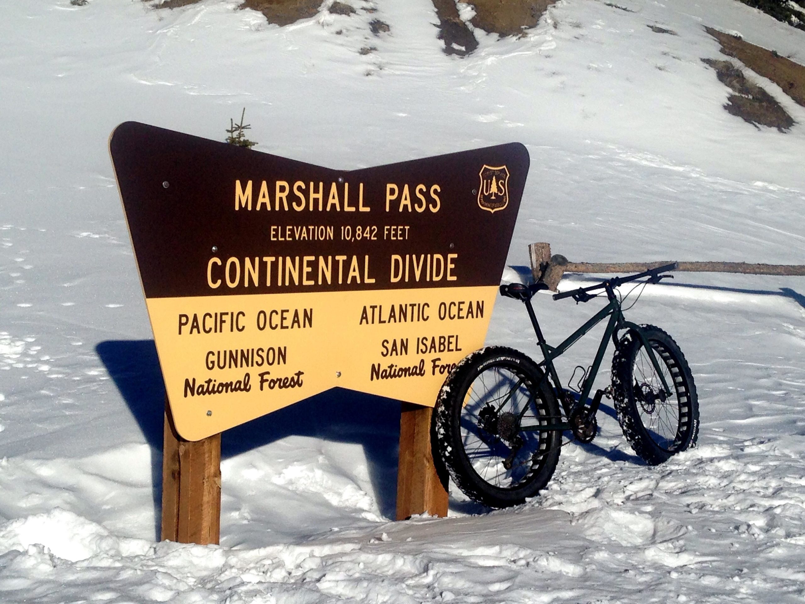 A brown and yellow sign indicating "Marshall Pass, Elevation 10,842 feet, Continental Divide" with directional information for the Pacific Ocean and Atlantic Ocean, alongside a fat tire mountain bike resting in the snow. The background features a snowy landscape with trees visible. Marshall Pass Road / #200 / #203 / #243 mountain bike trail.