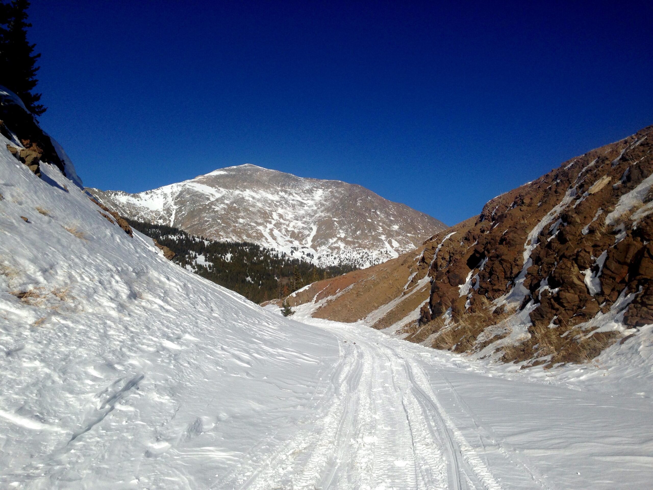 A snow-covered mountain trail flanked by rocky cliffs under a clear blue sky. The trail shows tire tracks leading towards a distant mountain peak, partially covered in snow, surrounded by evergreen trees. Marshall Pass Road / #200 / #203 / #243 mountain bike trail.