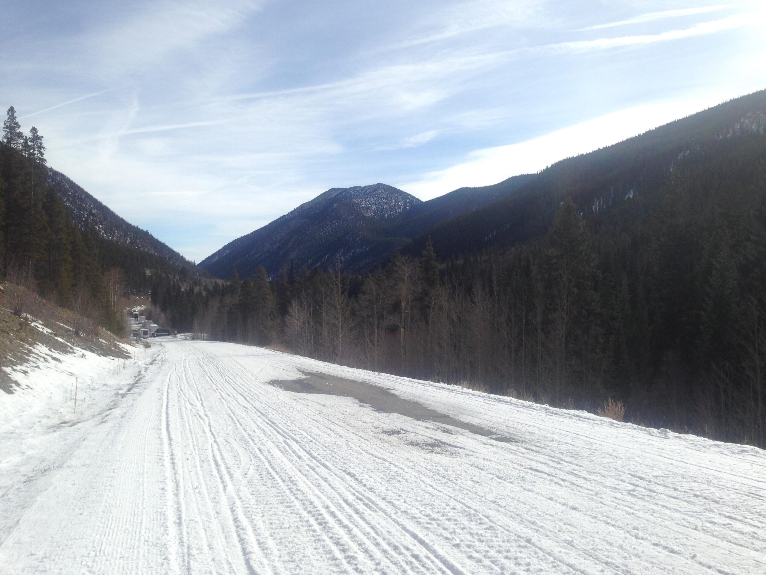 A snow-covered road winding through a valley surrounded by mountains. Pine trees line the sides, and the sky is partly cloudy with visible trails from airplanes. The scene captures a serene winter landscape. Cottonwood Pass Road mountain bike trail.