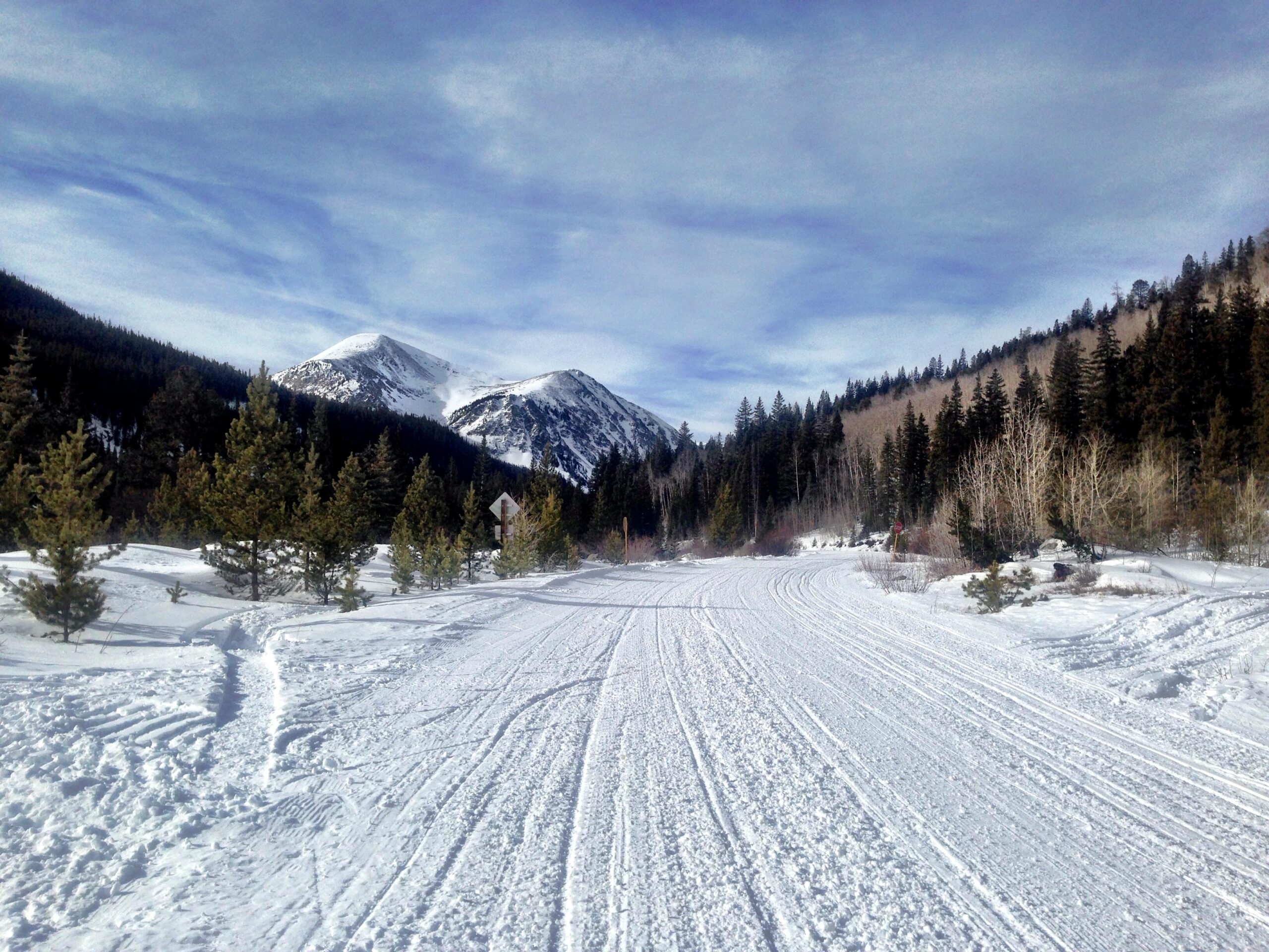 A snow-covered road winding through a mountainous landscape, flanked by evergreen trees and patches of snow. In the background, majestic peaks rise against a blue sky with wispy clouds, creating a serene winter scene. Cottonwood Pass Road mountain bike trail.