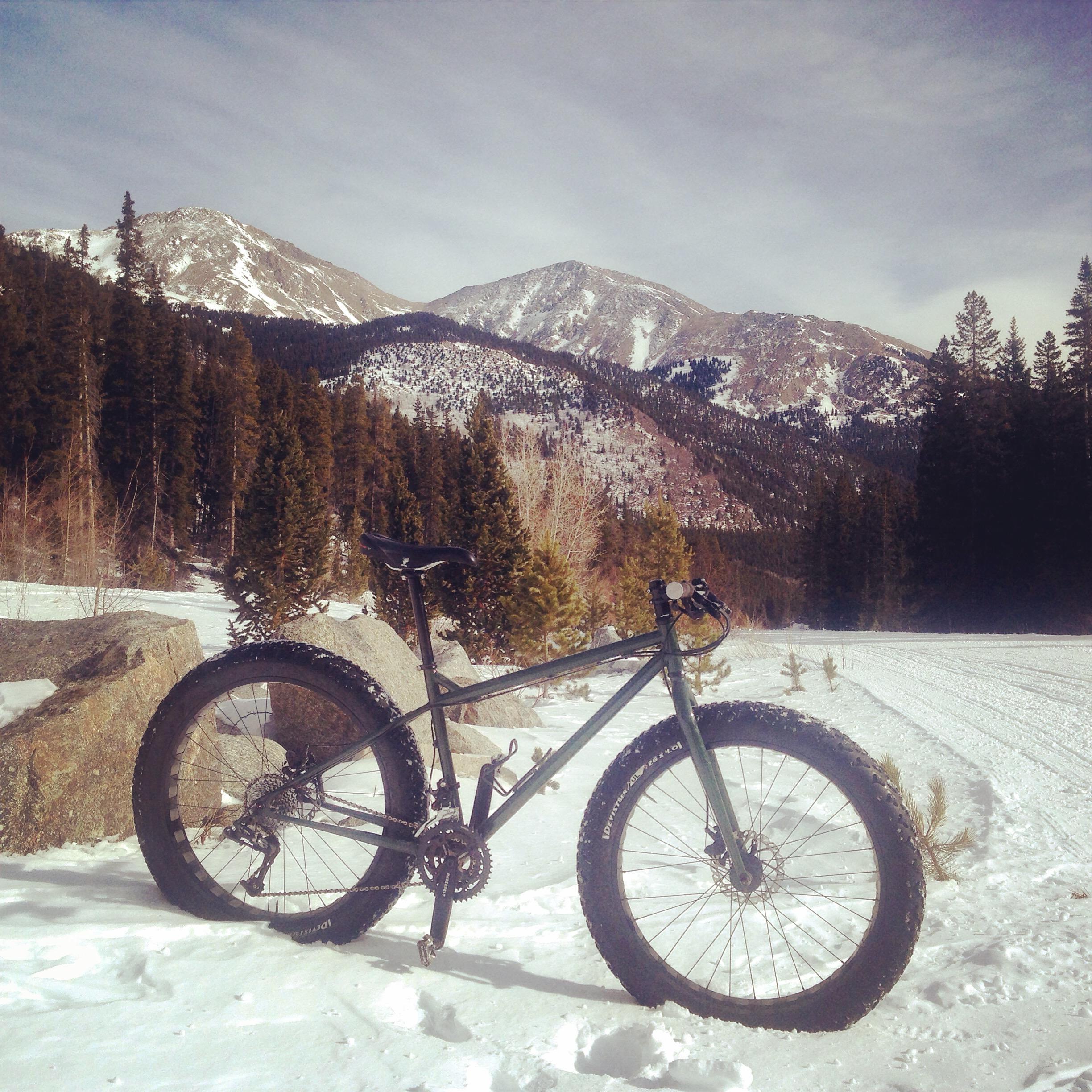A fat tire bike leaning against a rock, positioned in a snowy landscape with tall pine trees, mountains in the background, and a partly cloudy sky. Cottonwood Pass Road mountain bike trail.