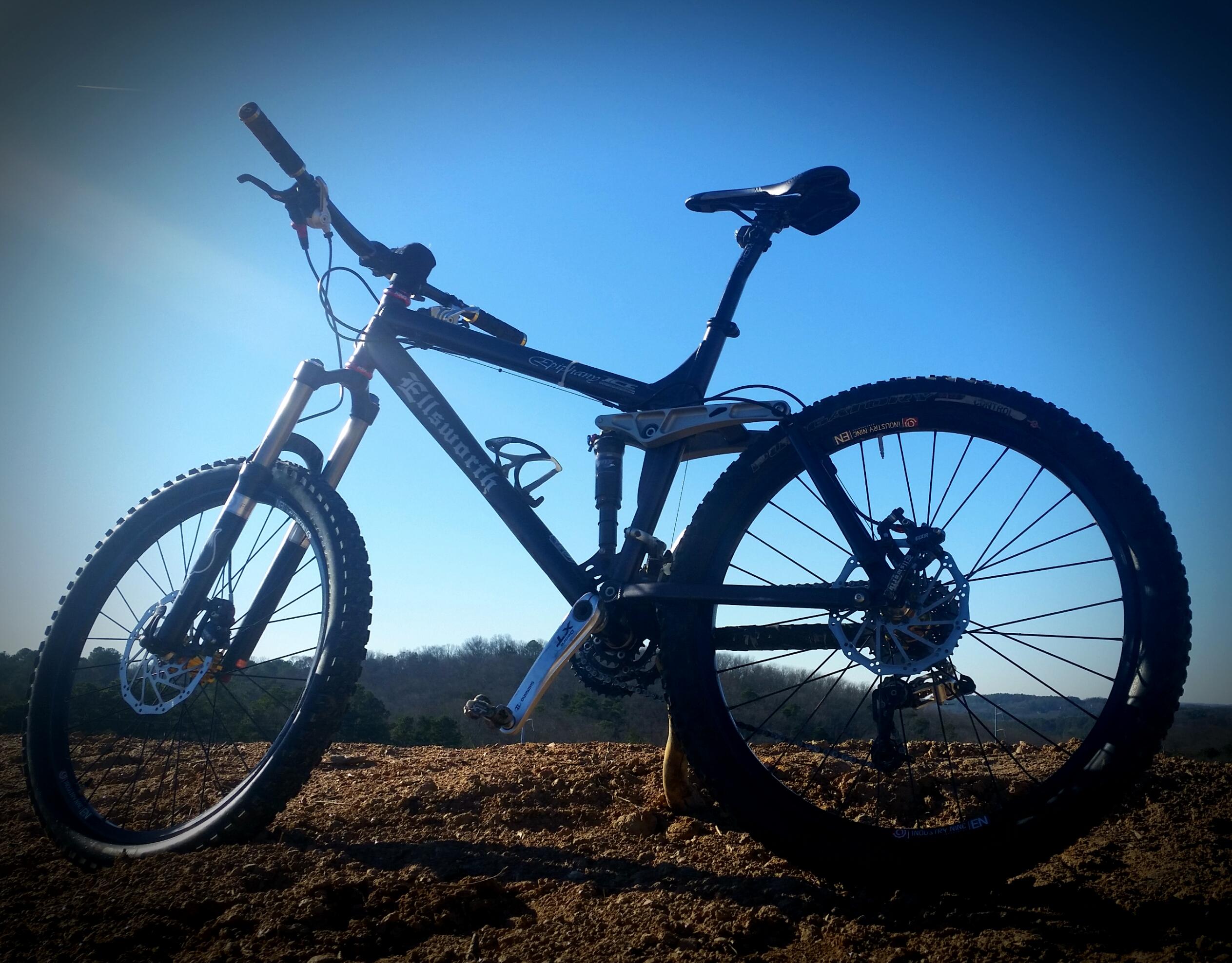 Ellsworth Epiphany: A mountain bike positioned on a dirt surface with a clear blue sky in the background. The bike features sturdy tires and dual suspension, showcasing its design for off-road riding. The image highlights the bike's frame and components, emphasizing its sporty appearance.
