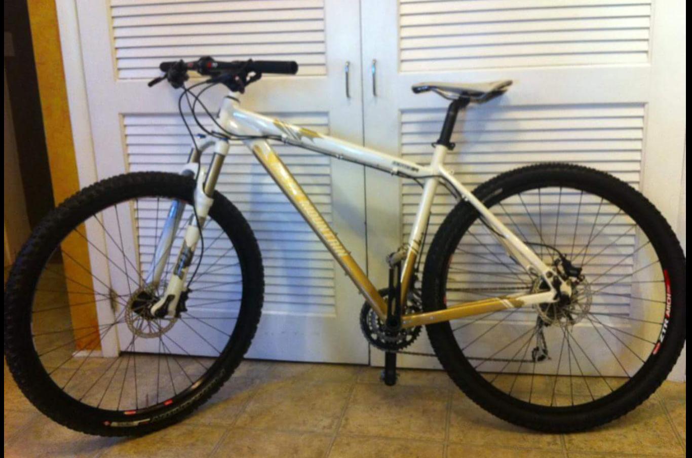 Rocky Mountain Vertex 990 rsl 2013: Image of a mountain bike with a white and gold frame, featuring thick, knobby tires. The bike stands upright against a backdrop of closed louvered doors. The handlebars are equipped with grip and brake levers, while the bike's drivetrain is visible near the rear wheel. The flooring is tiled.