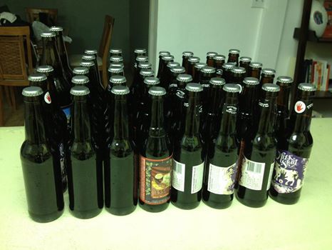 A collection of approximately 50 dark glass bottles arranged on a countertop, some with colorful labels, in a well-lit indoor space.