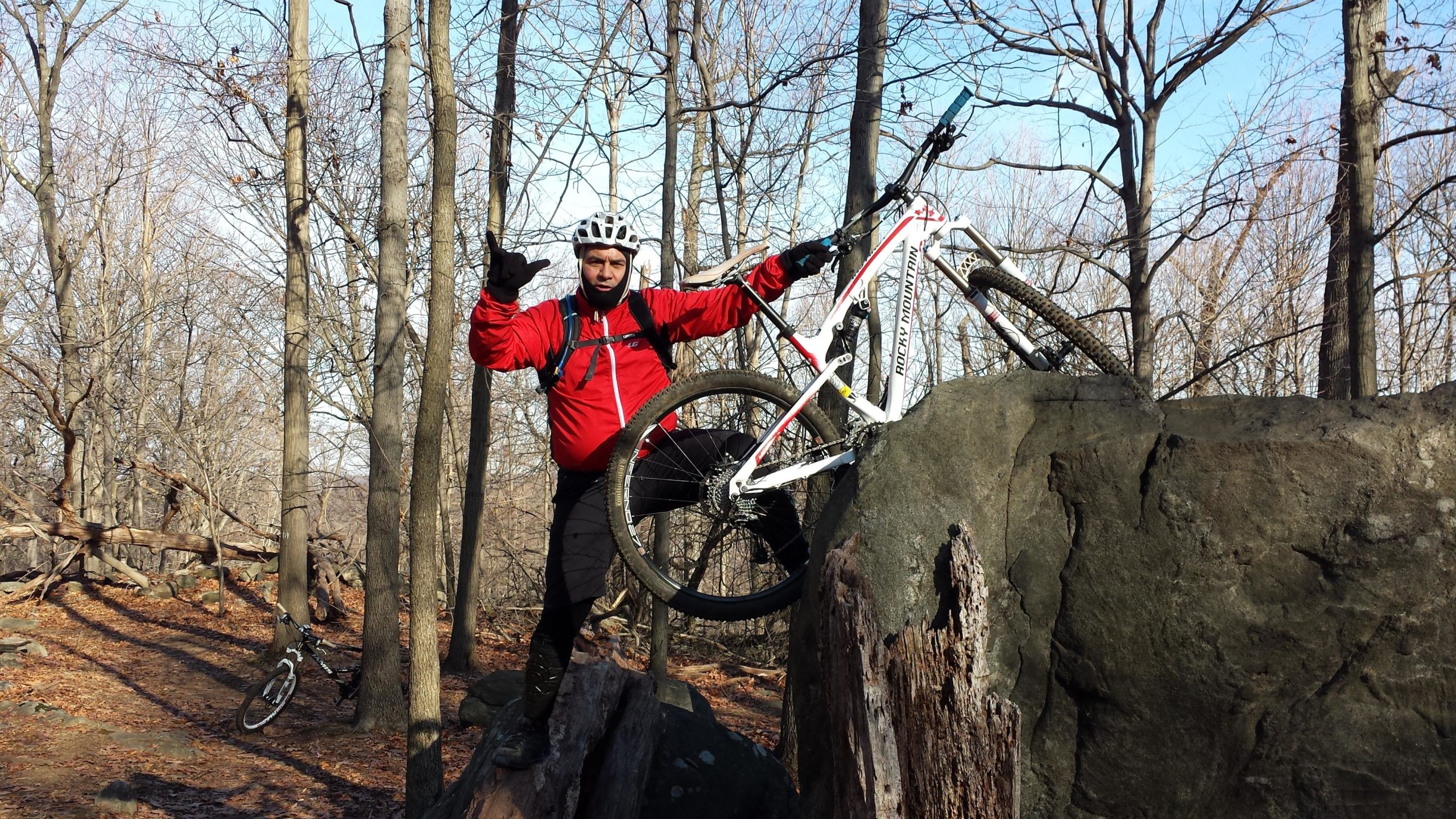 A person wearing a red jacket and a helmet stands confidently on top of a large rock, one foot raised on the rock and the other on the ground, while holding a mountain bike next to them. The background features a wooded area with leafless trees and fallen branches, typical of an outdoor trail. The individual is making a hand gesture, appearing to enjoy the moment in nature. Sprain Ridge mountain bike trail.