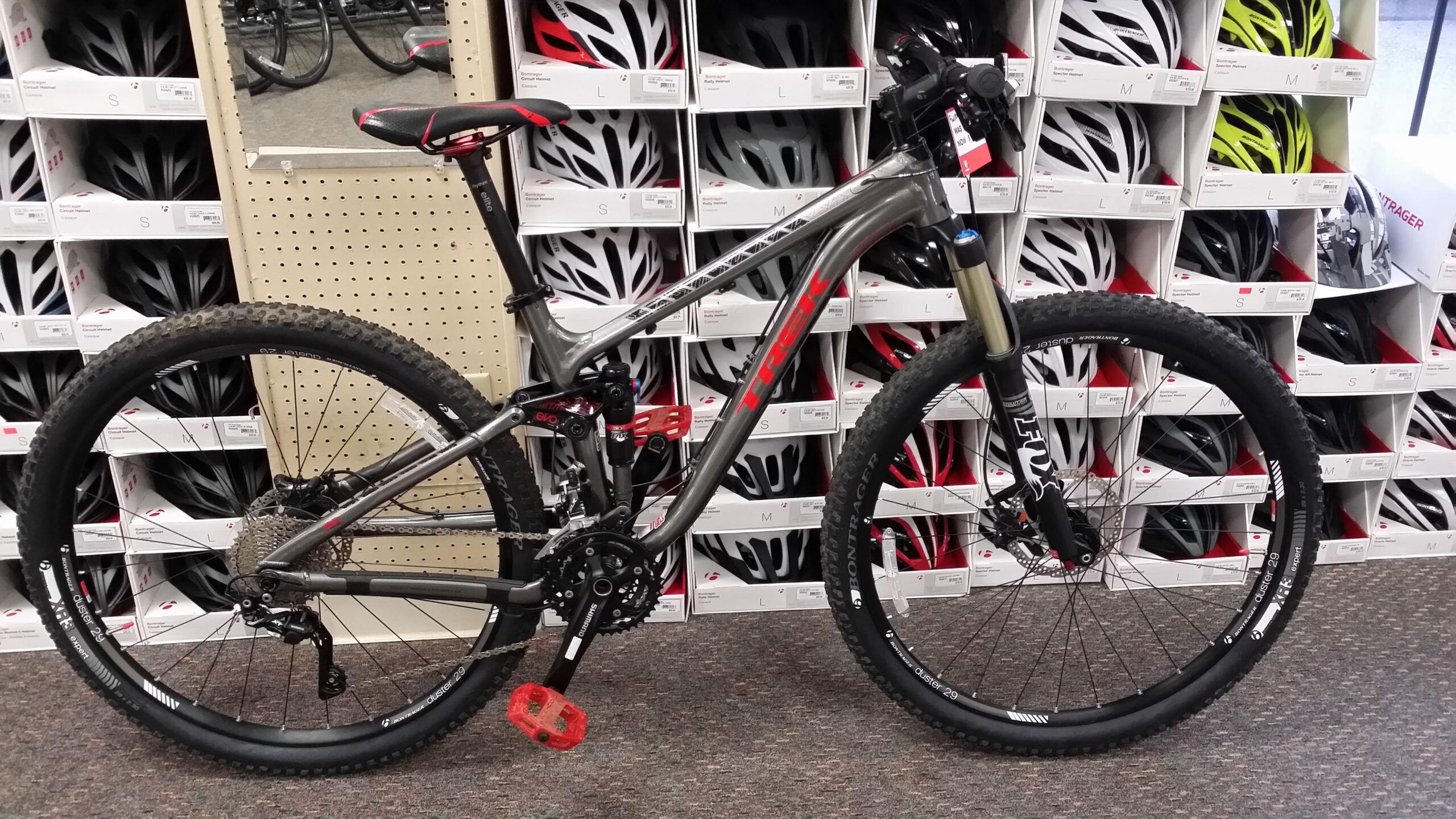 Trek Fuel EX 8 29: A mountain bike with a silver frame and red accents is displayed in a store. The bike features thick, knobby tires suitable for off-road riding, a front suspension fork, and red platform pedals. In the background, neatly arranged bicycle helmets in various sizes are visible on shelves.