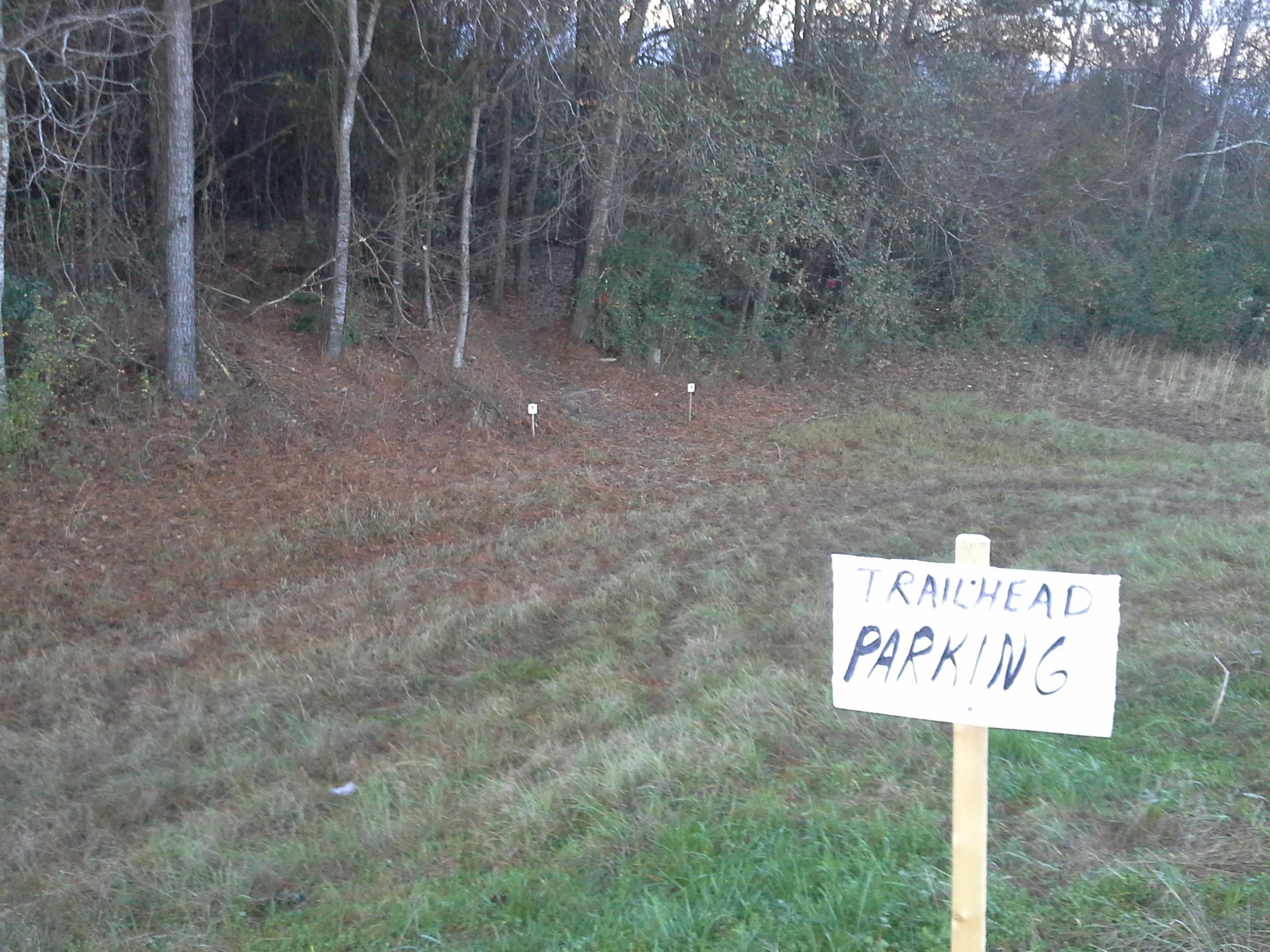 A handwritten sign that reads "TRAILHEAD PARKING" is displayed on a wooden post in the foreground, with a grassy area and trees in the background. The scene suggests the location of a parking area near a hiking trail, surrounded by foliage and a dirt path leading into the woods. Petal River Park mountain bike trail.