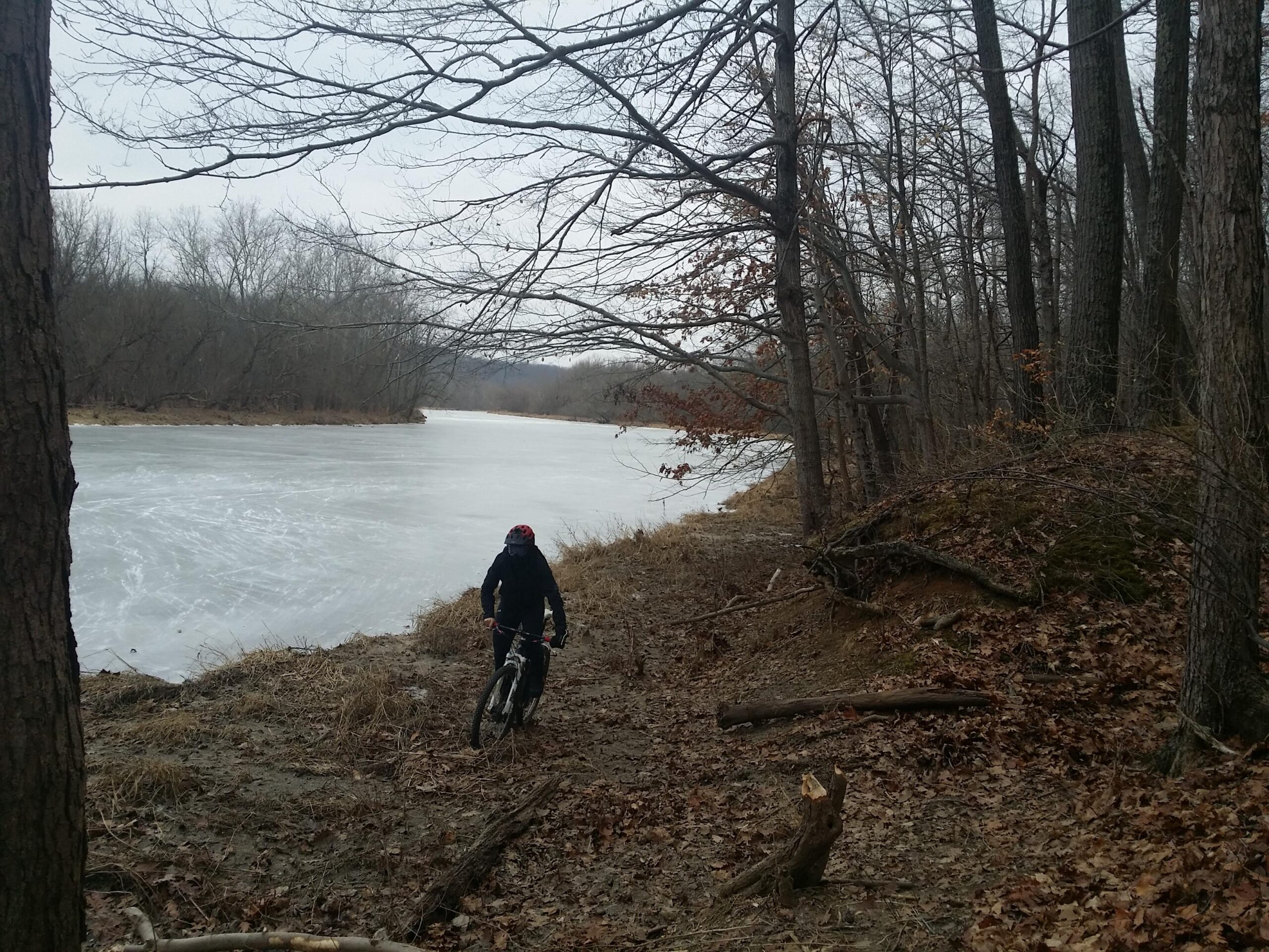 A person riding a mountain bike along a gravel path beside a frozen river, surrounded by bare trees and autumn leaves, on a cloudy day. Versailles State Park mountain bike trail.