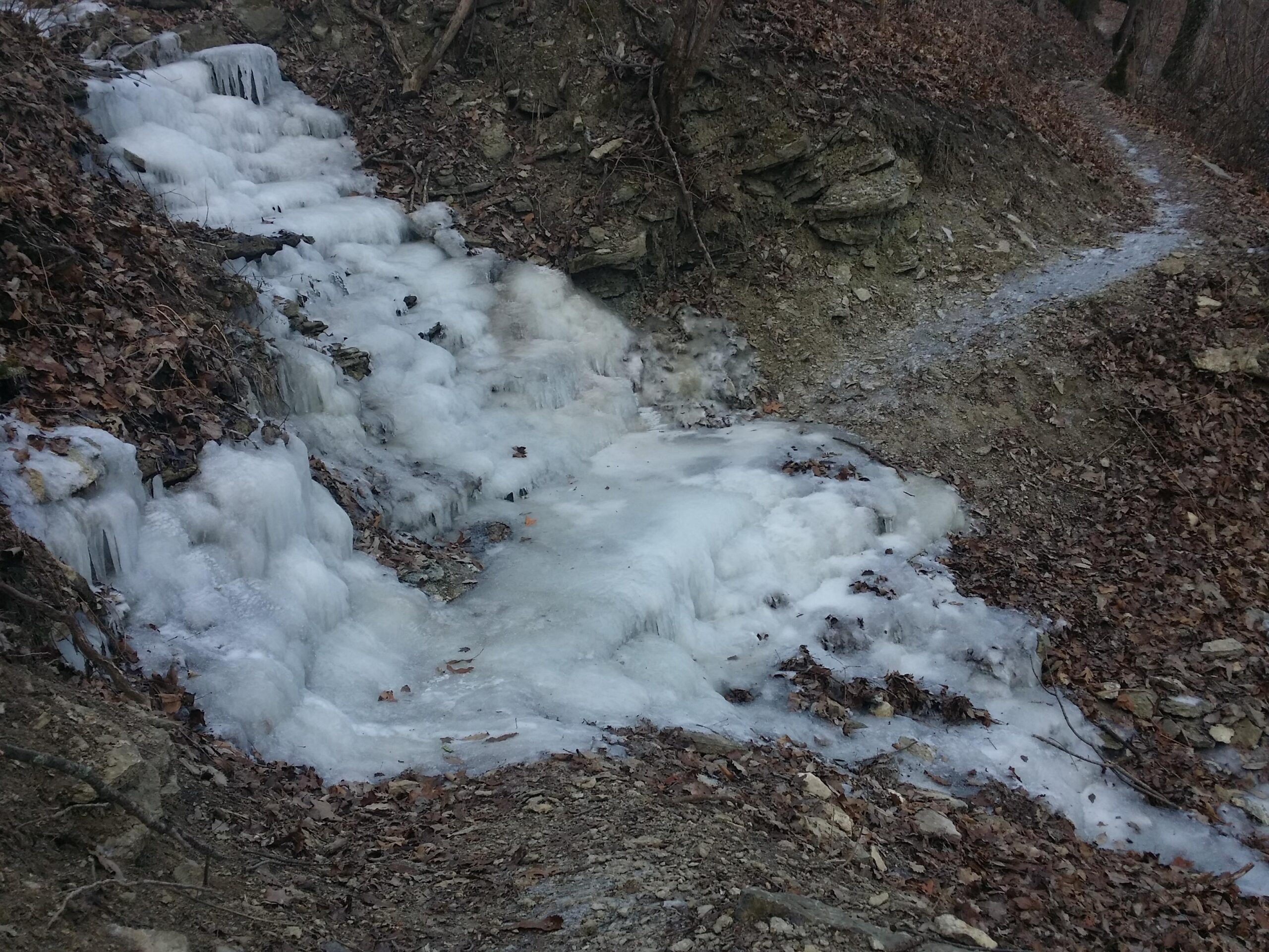 A frozen waterfall cascading down a rocky slope, surrounded by dry leaves and bare trees. A narrow, winding path runs alongside the icy terrain, indicating a trail through a winter landscape. Versailles State Park mountain bike trail.
