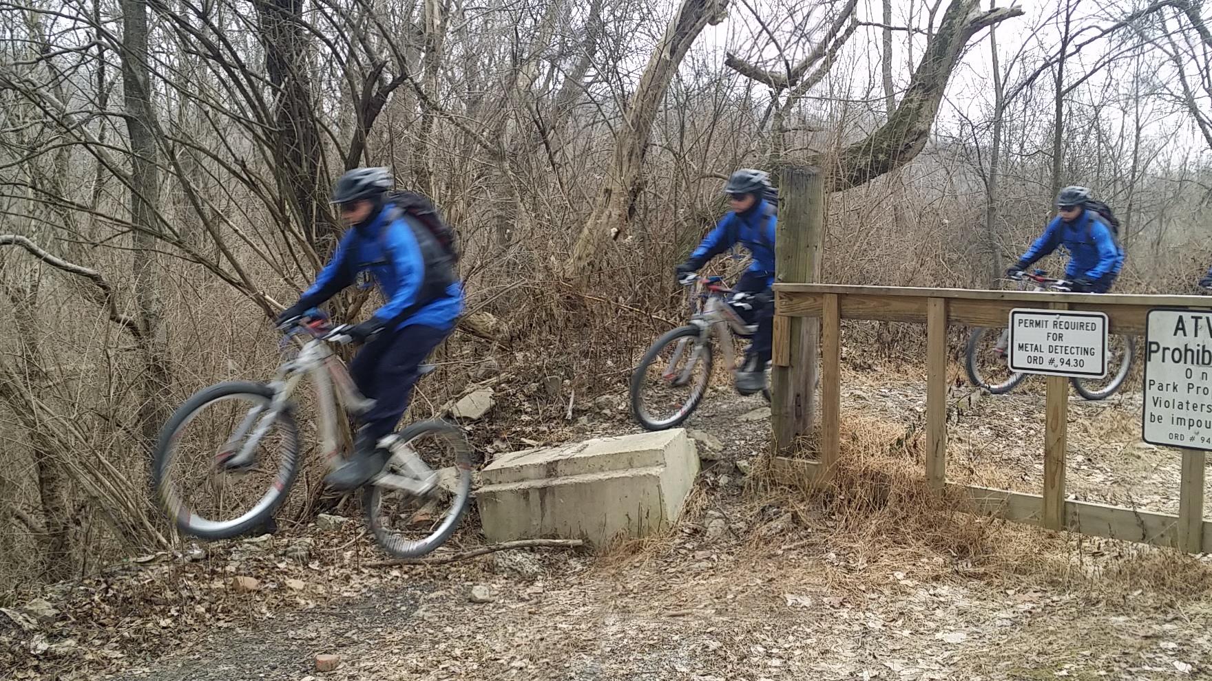 A cyclist wearing a blue jacket and helmet rides a mountain bike on a gravel path surrounded by bare trees and brush. The image shows the cyclist in motion, with a wooden barrier and a sign indicating permit requirements visible in the background. Devou Park mountain bike trail.
