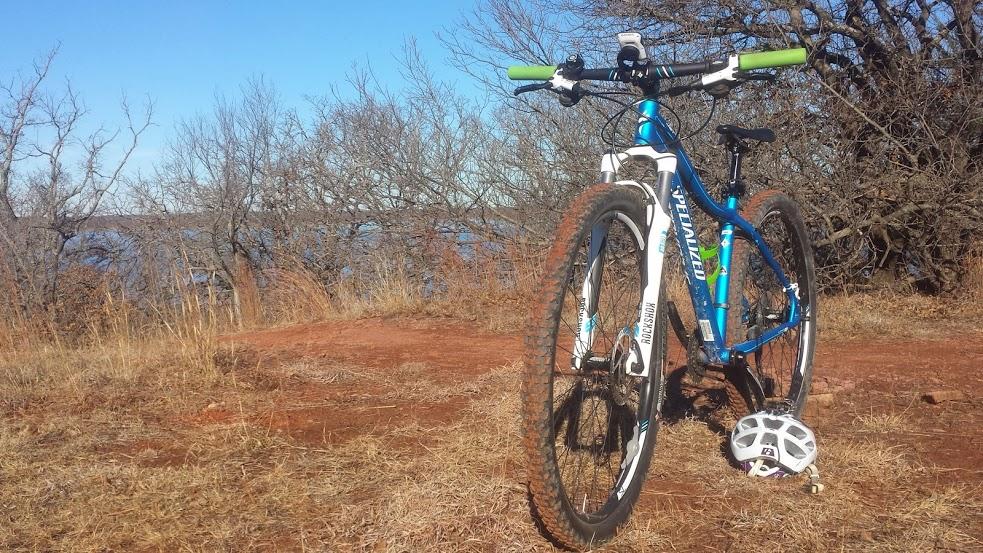 A blue mountain bike with green handlebars is parked on a dirt path. In the background, there are bare trees and a view of a lake under a clear blue sky. A white helmet is resting on the ground next to the bike. Thunderbird Lake Clear Bay mountain bike trail.