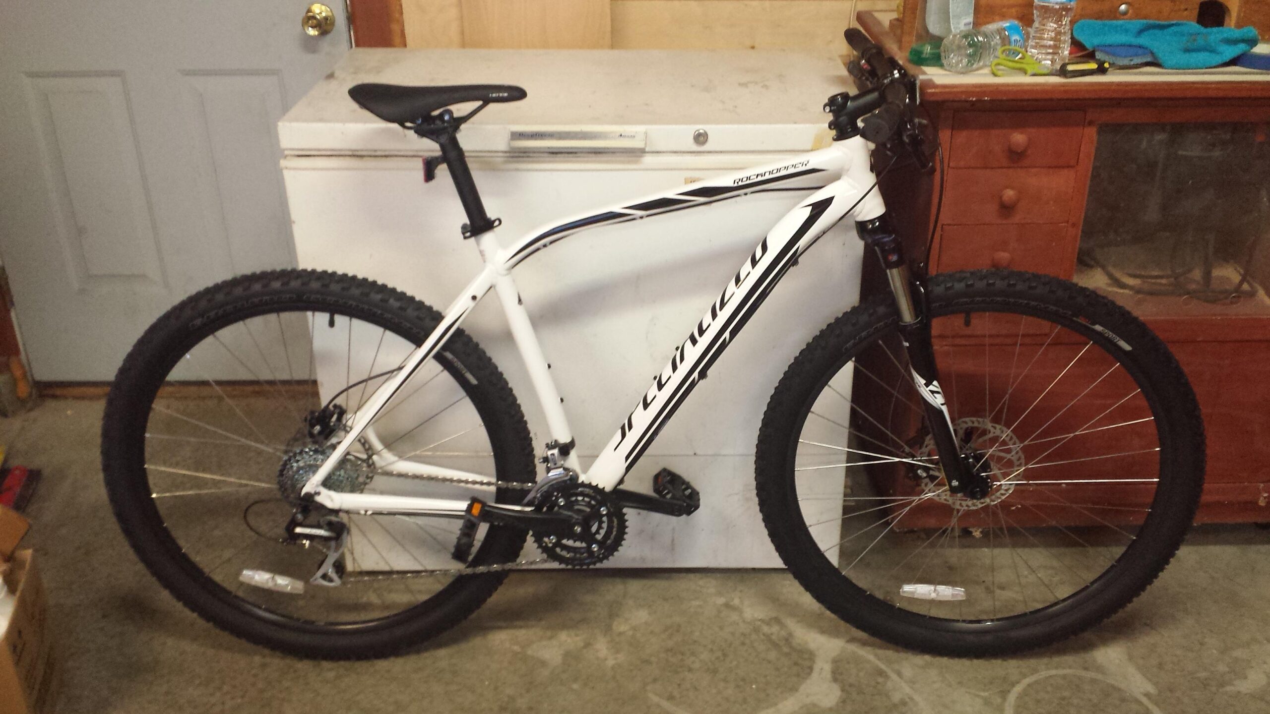 Specialized Rockhopper 29: A white mountain bike is leaning against a wall in a garage setting. The bike features wide tires, front suspension, and a black saddle. In the background, there is a white chest freezer and a wooden cabinet with various items, including a water bottle and tools. The garage floor is concrete with some scattered boxes.