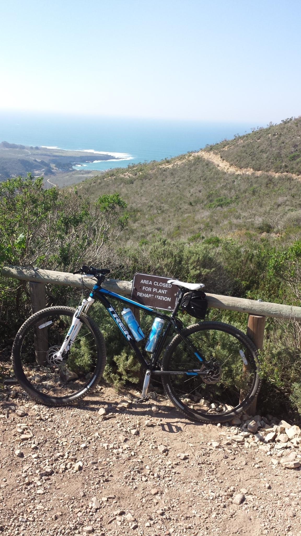 Trek EX 8: A mountain bike leaning against a wooden fence with a sign that reads "Area Closed for Plant Rehabilitation." In the background, there is a scenic view of a coastline with the ocean and hilly terrain, surrounded by greenery and shrubs under a clear sky. The ground is rocky and sandy, typical of a trail environment.