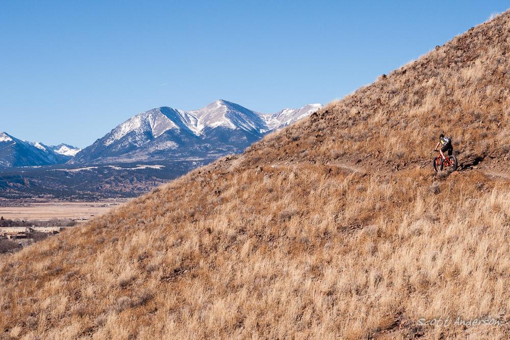 A cyclist riding along a narrow dirt path on a hillside, with dry grasses and rocky terrain in the foreground. The background features snow-capped mountains under a clear blue sky, highlighting a stunning outdoor landscape. Arkansas Hills mountain bike trail.