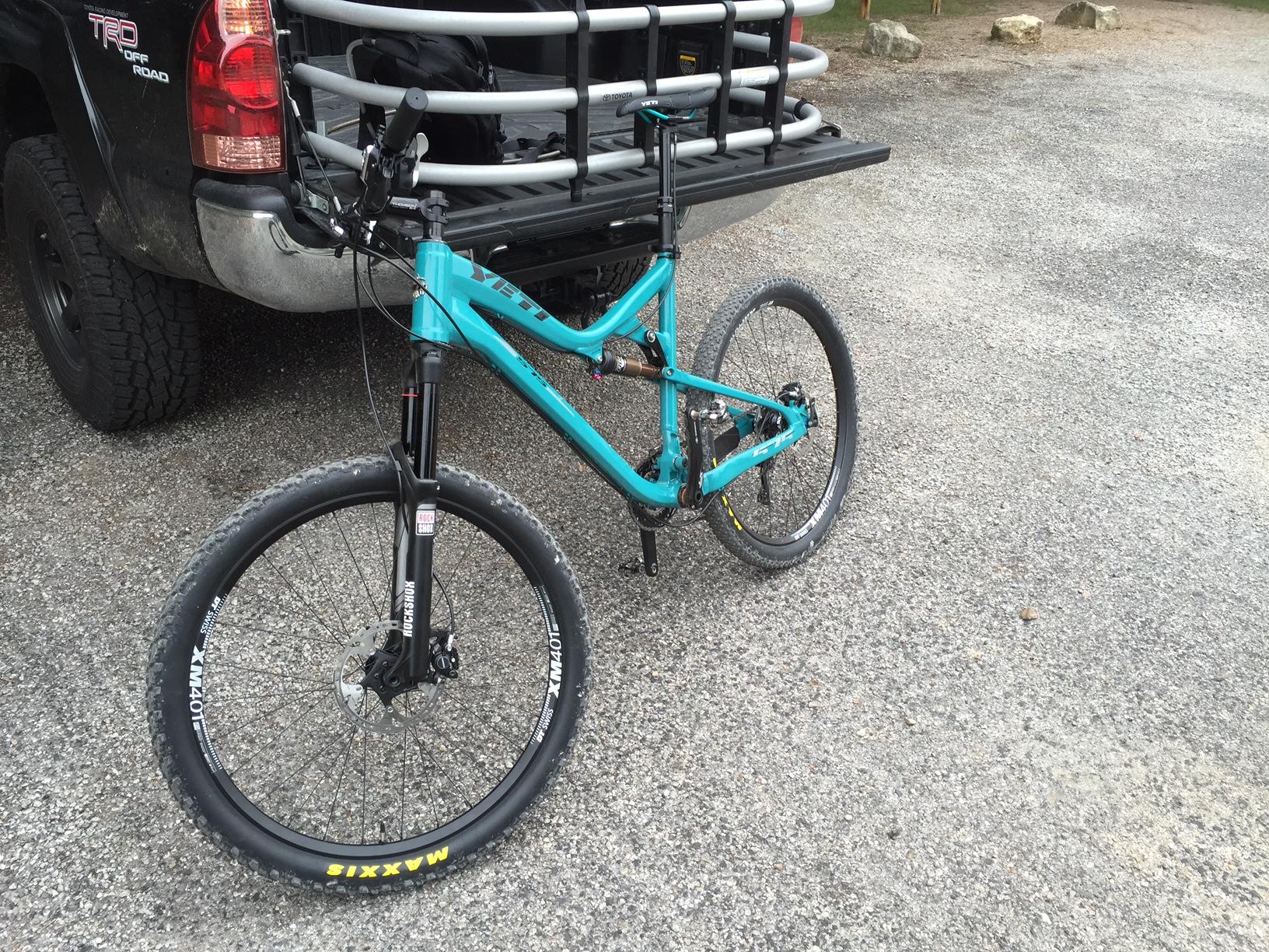Yeti 575: A turquoise mountain bike parked on gravel next to a pickup truck with a bike rack. The truck has a black finish and displays 'TRD Off Road' on the tailgate. The bike features thick tires and a front suspension fork.