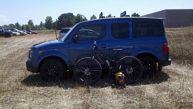 Specialized Hardrock Disc 29: A blue SUV parked in an open field alongside a black mountain bike and a yellow backpack. In the background, there are other parked cars and trees lining the horizon. The scene is sunny with dry grass visible in the foreground.