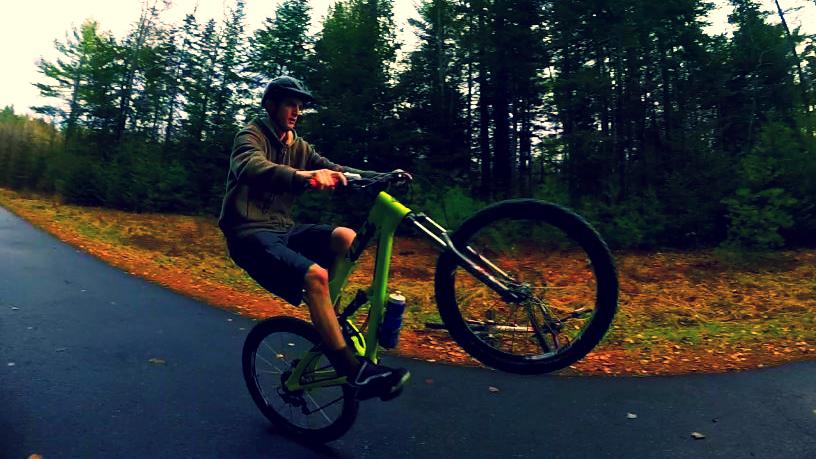 Yeti SB66: A person performing a wheelie on a bright green mountain bike on a forested pathway, surrounded by trees and fallen leaves. The rider is dressed in a hoodie and shorts, with a dark cap, showcasing a dynamic action pose mid-ride.