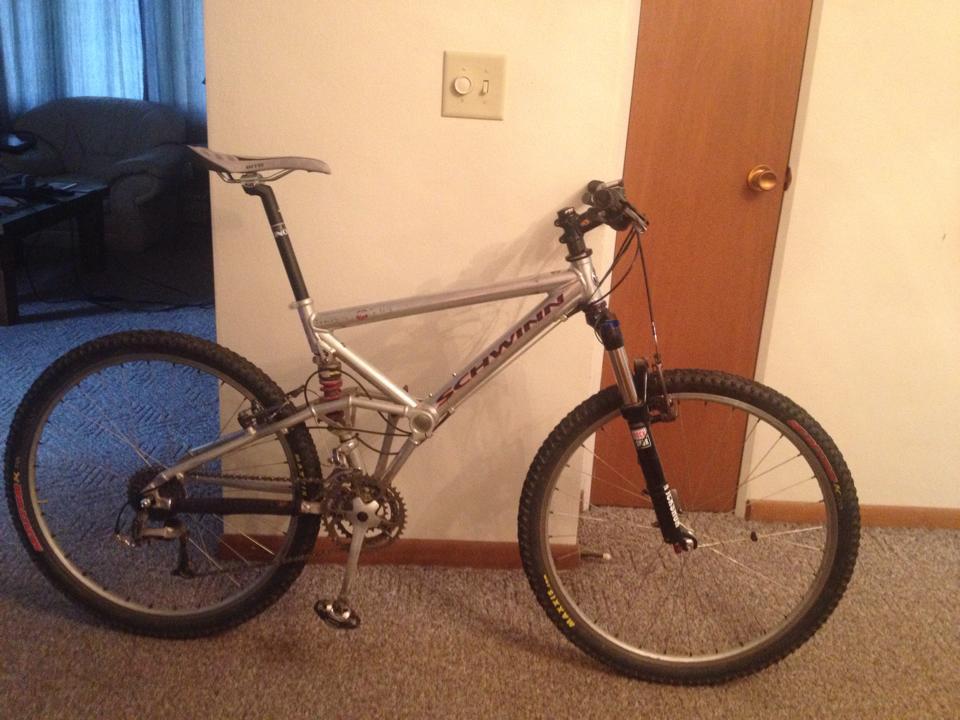 Schwinn Homegrown: A silver Schwinn mountain bike is leaning against a wall in a living room. The bike features thick tires and a front suspension fork, with a visible gear system and handlebars. In the background, a sofa and a wooden door are partially visible. The floor is covered with carpet.
