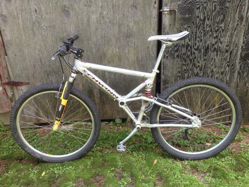 Schwinn Homegrown: A silver Schwinn mountain bike with front suspension, resting on green grass in front of a weathered wooden fence. The bike features knobby tires and a white saddle, positioned upright for display.