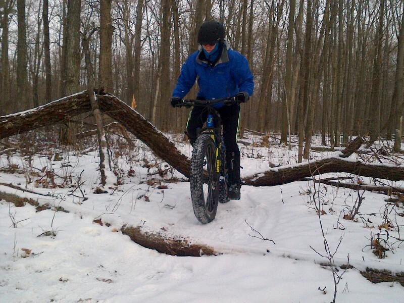 A person riding a fat bike navigates over a fallen log in a snowy forest, wearing a blue jacket and helmet, surrounded by bare trees. Dufferin County mountain bike trail.
