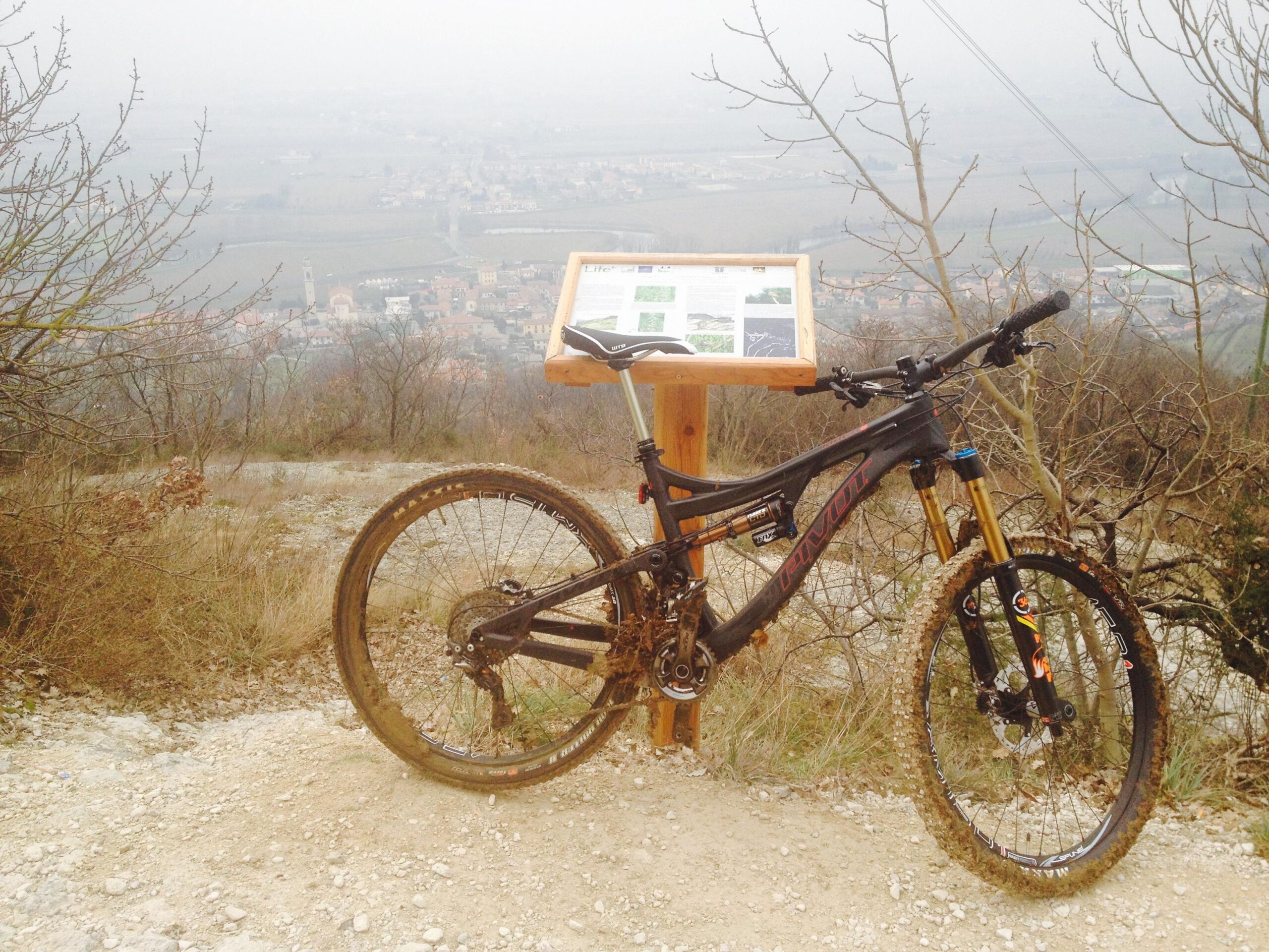 Pivot Mach 6: Mountain bike resting on a trail near a viewpoint, with a wooden information sign in the background. The landscape features a hazy view of a town below, surrounded by fields and trees, indicating an outdoor recreational area. The bike has mud on the tires, suggesting recent use on a rugged path.