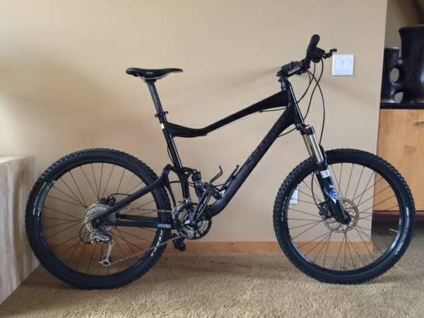 Giant Trance X2: A black mountain bike with thick tires and a front suspension, positioned against a beige wall. The bike features a sleek frame design and is equipped with multiple gears and a disc brake system. The background includes a wooden floor and a piece of furniture.