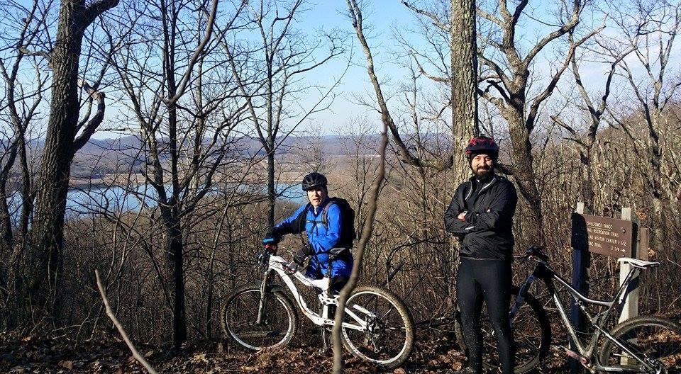 Two mountain bikers pause for a photo while standing on a scenic overlook among bare trees. In the background, a lake is visible, surrounded by hills under a clear blue sky. One biker is wearing a blue jacket and helmet while the other is dressed in a black outfit. Both bikes are parked nearby. A sign indicating the trail is also present in the image. Cave-run Lake mountain bike trail.