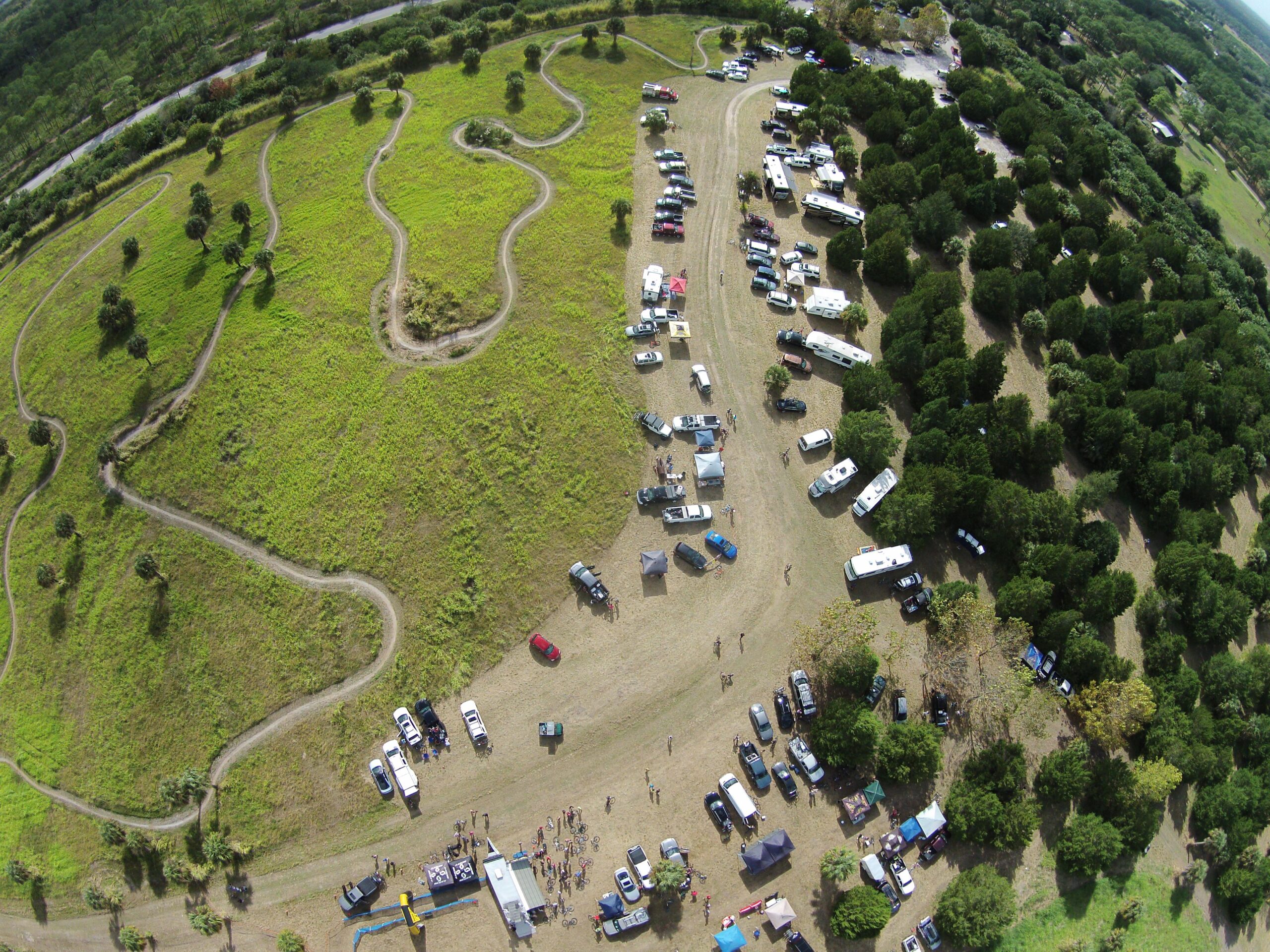 Aerial view of a grassy area with winding dirt paths, surrounded by trees. Multiple vehicles and tents are parked on the ground, with people gathered in various locations. A road is visible in the background, indicating the setting is likely a community event or gathering. Caloosahatchee Regional Park mountain bike trail.