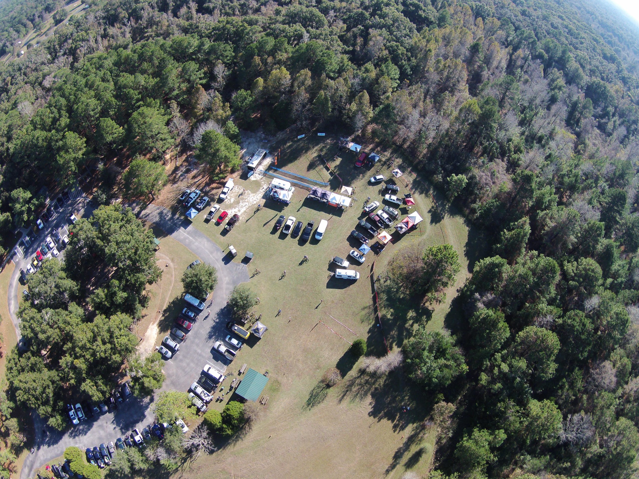 Aerial view of a wooded area showing a parking lot filled with vehicles. In the foreground, there are several tents and outdoor setups, with people scattered around. The surrounding landscape is lush with trees, indicating a natural setting. Santos mountain bike trail.