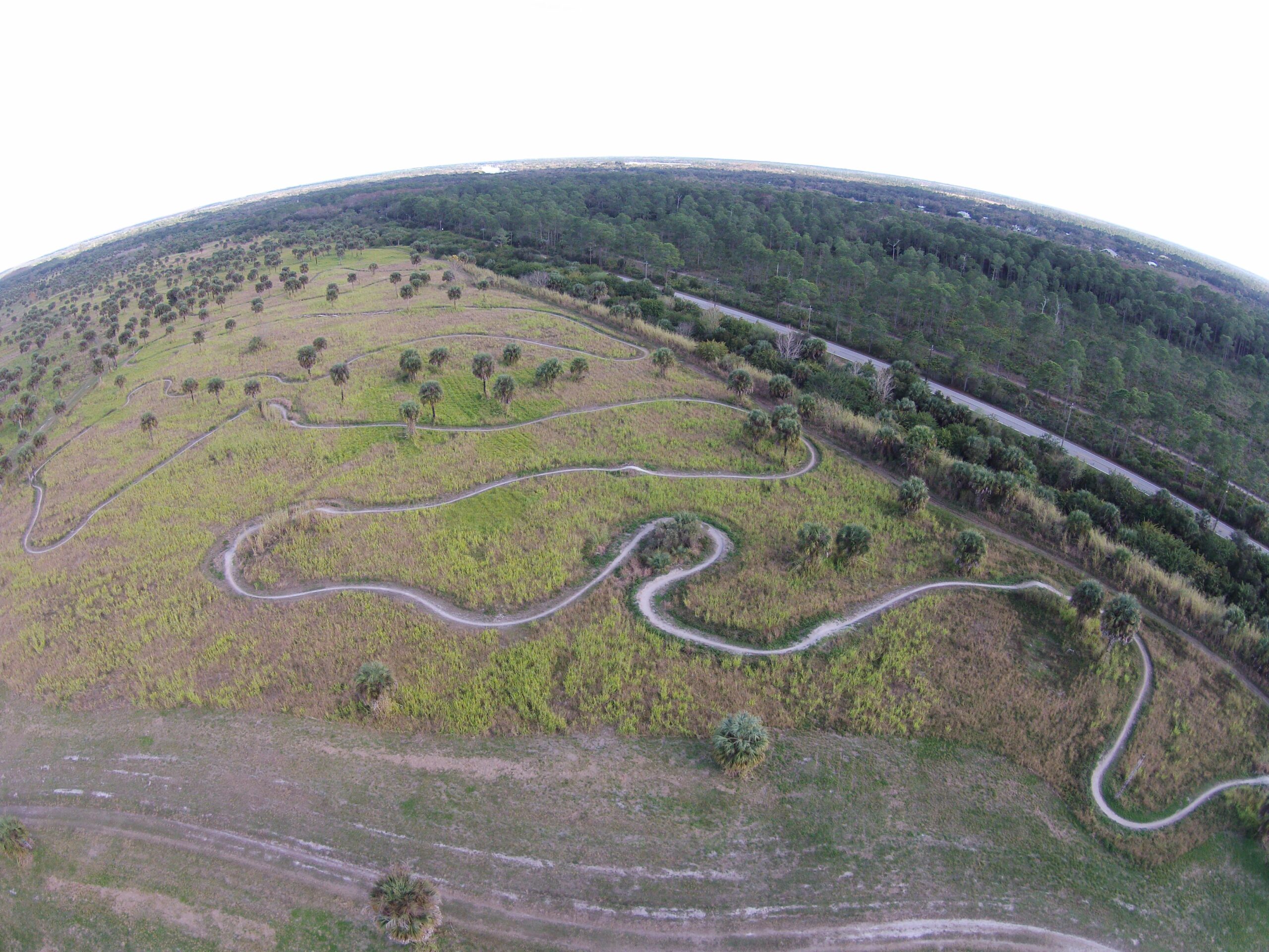 Aerial view of a winding trail through a grassy landscape dotted with small trees, surrounded by dense forests in the background. The scene captures a mixture of green and brown hues of the land. Caloosahatchee Regional Park mountain bike trail.