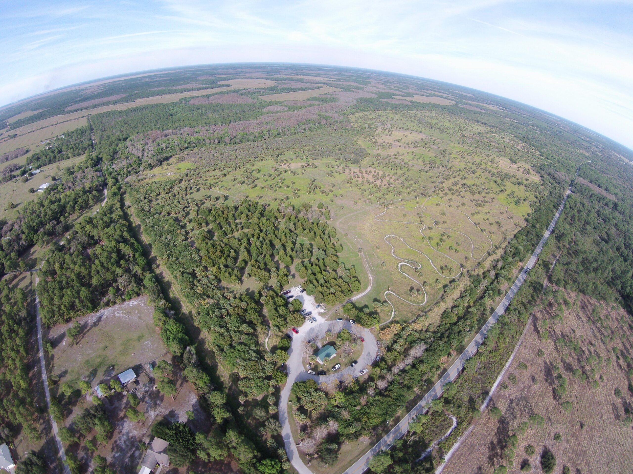 Aerial view of a sprawling landscape featuring dense greenery and winding roads. The image showcases a mix of wooded areas and open fields under a clear blue sky. A small cluster of buildings and parked cars can be seen near the bottom of the image. Caloosahatchee Regional Park mountain bike trail.