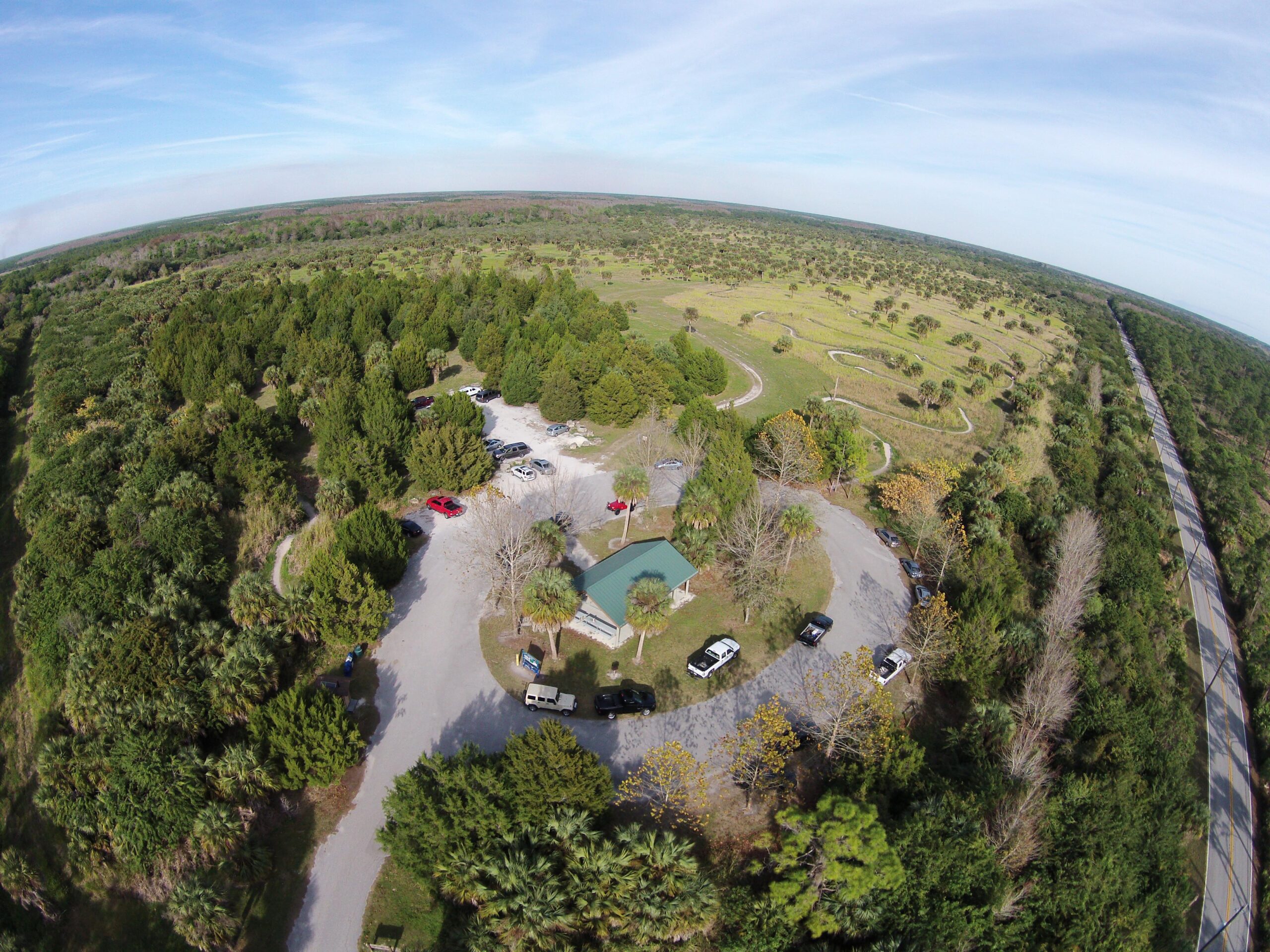 Aerial view of a wooded area with a parking lot and a small building. The landscape features a mix of trees and open fields, along with winding paths. A road runs alongside the edge of the area, suggesting access to the location. The sky is clear with a few clouds visible. Caloosahatchee Regional Park mountain bike trail.