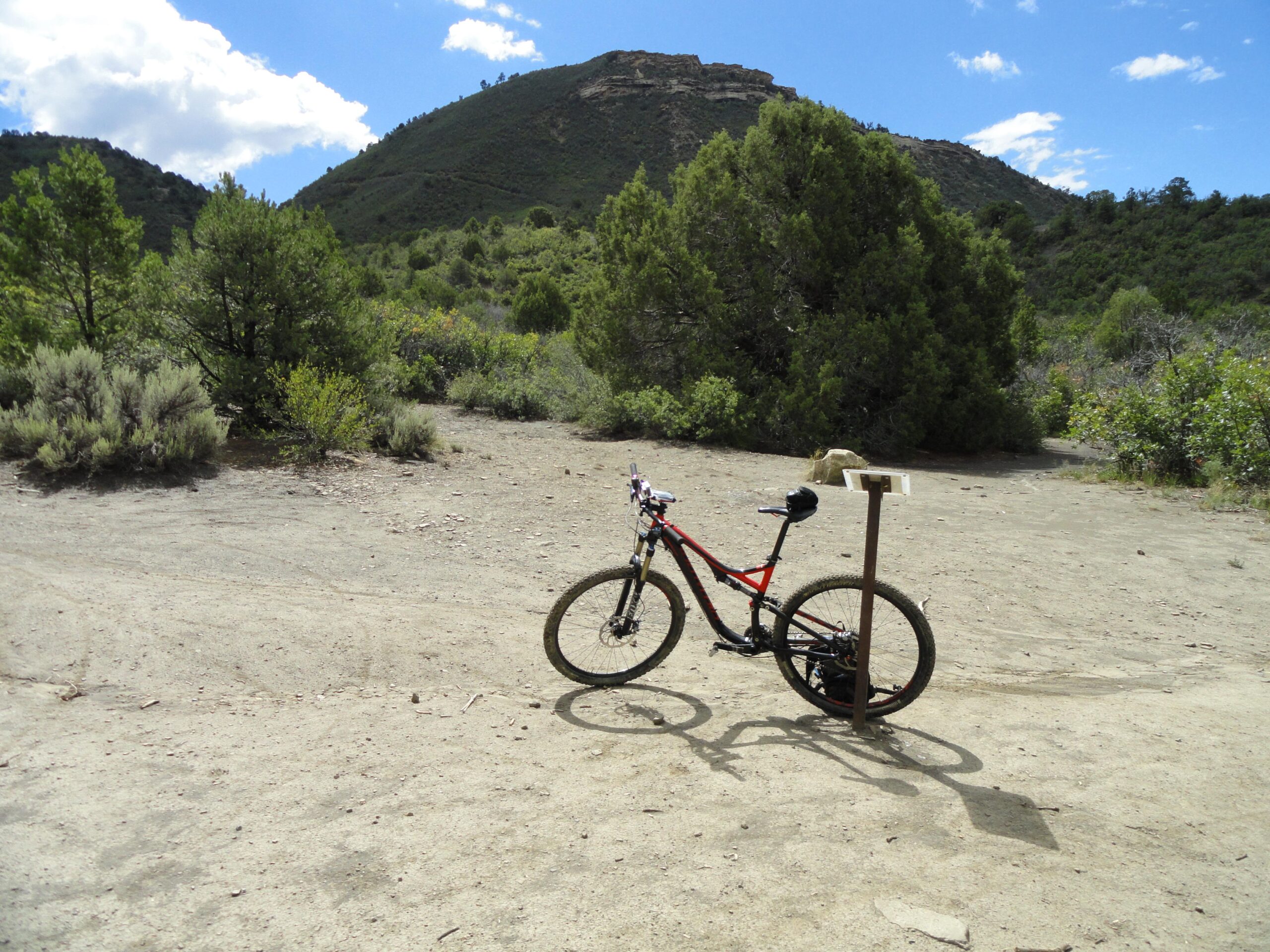Specialized Stumpjumper FSR Comp 29: A mountain bike is parked next to a signpost on a dirt trail surrounded by greenery, with a hilly landscape in the background and a partly cloudy blue sky.