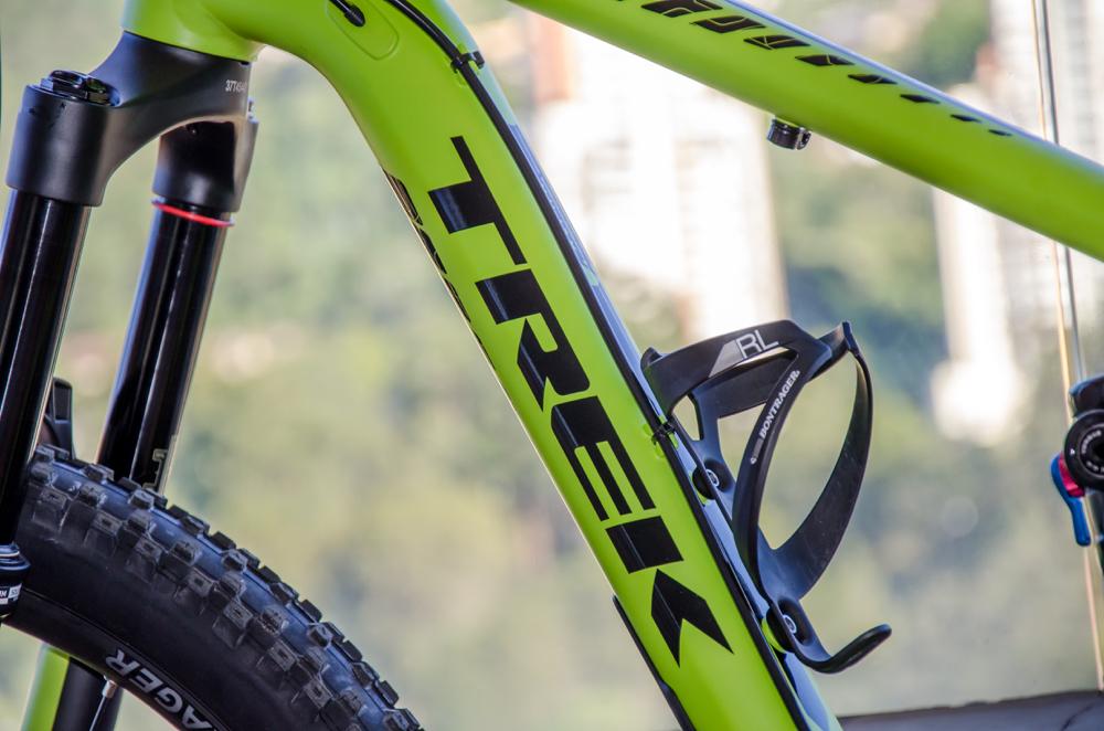 Trek Remedy 7 27.5: Close-up of a bright green Trek bicycle frame featuring a black water bottle holder and front suspension fork. The background shows a blurred outdoor landscape, suggesting a city or natural environment.