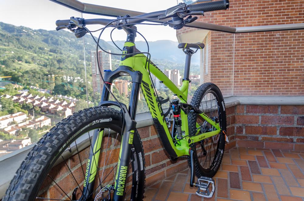 Trek Remedy 7 27.5: A brightly colored green mountain bike leaning against a balcony railing, with a scenic view of a mountainous landscape and residential buildings in the background. The bike features large, rugged tires and a modern frame design.