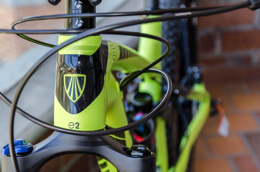 Trek Remedy 7 27.5: Close-up view of a vibrant green bicycle frame, featuring a shield emblem on the front with the text "e2." The image showcases intertwined cables and components, with a blurred background of brick and tile, highlighting the bike's design.