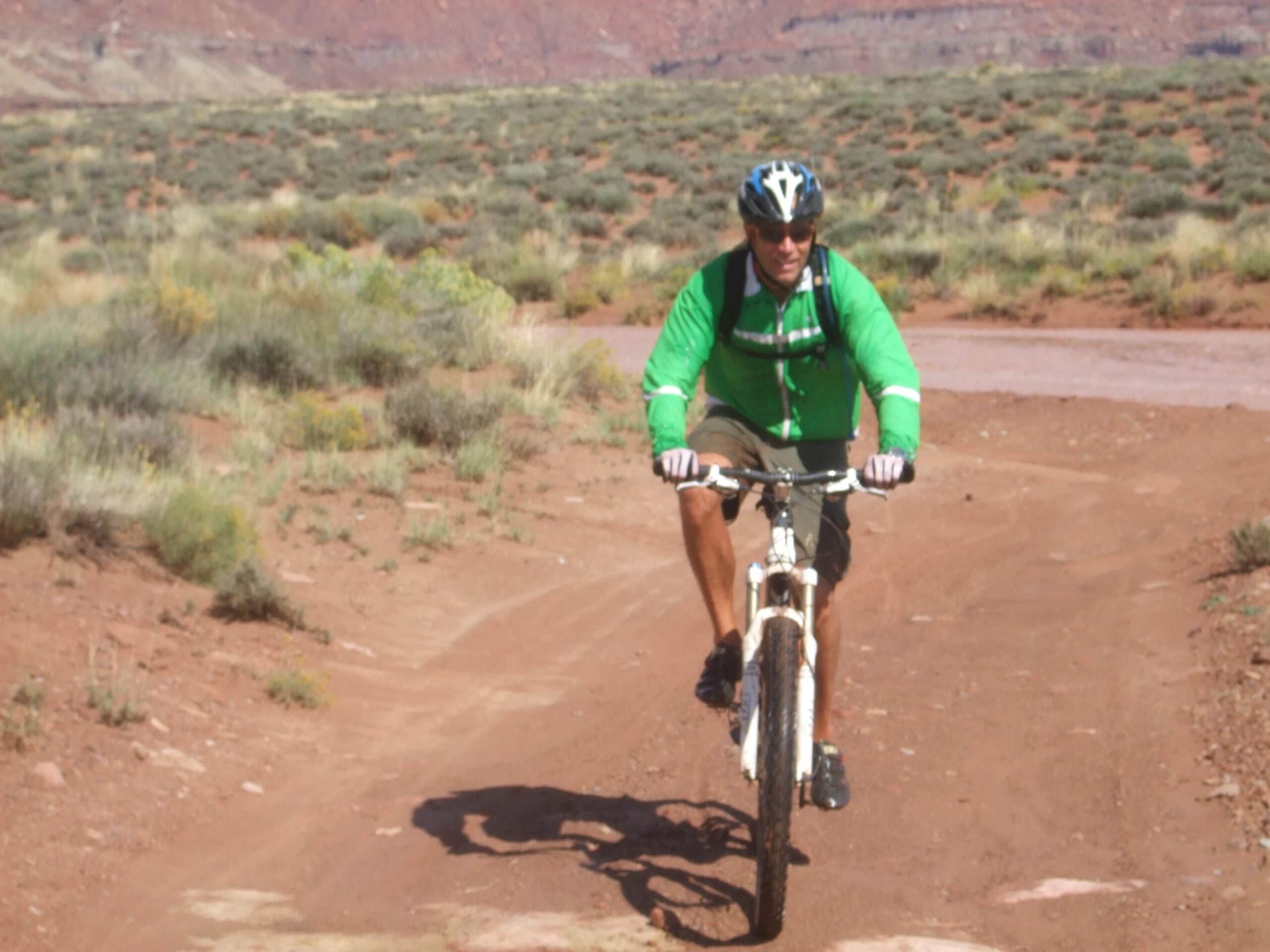 Trek Superfly 100 Elite carbon: A person wearing a green jacket and a helmet is riding a mountain bike along a dirt path in a natural landscape. The background features sparse vegetation and rocky terrain, typical of a desert environment.