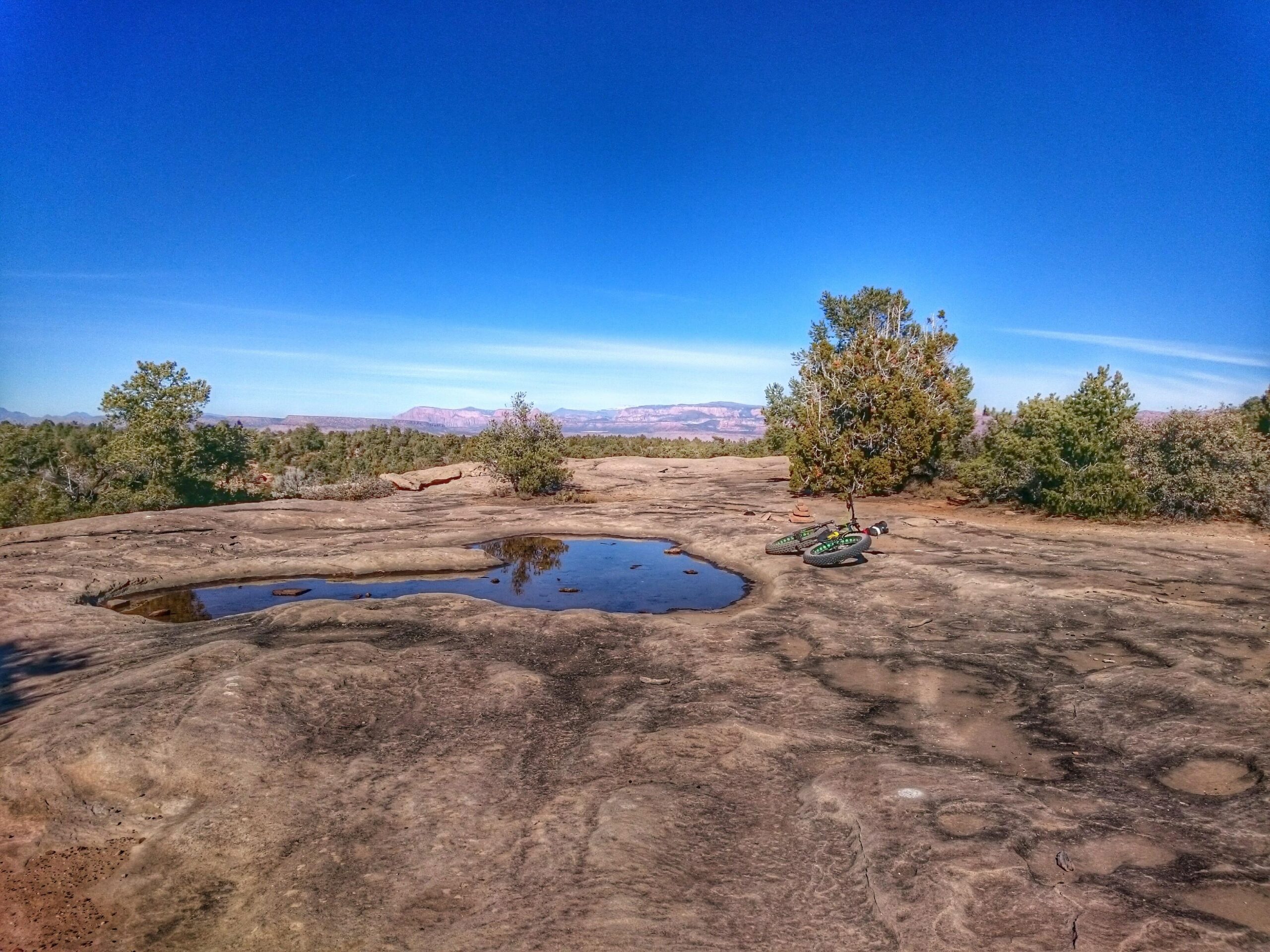 A rocky landscape under a clear blue sky, featuring a large puddle reflecting the surroundings. A mountain bike leans against a sparse tree, surrounded by shrubs and distant mountains. The terrain shows distinct rock formations with visible textures. Gooseberry Mesa mountain bike trail.