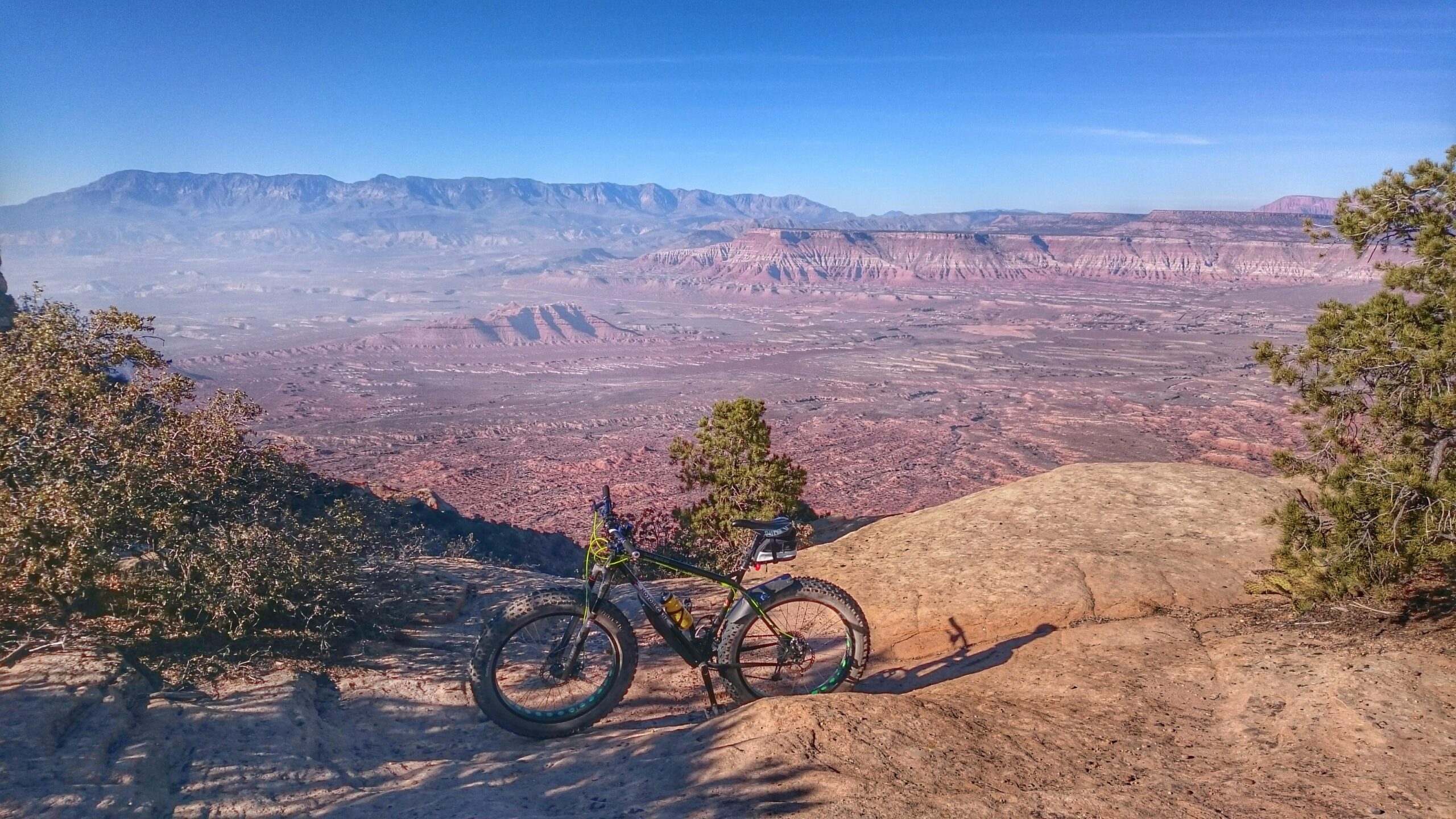 A mountain bike resting on a rocky surface with a panoramic view of a desert landscape, featuring layered hills and distant mountains against a clear blue sky. Vegetation is visible in the foreground, adding contrast to the vast expanse of the scenery. Gooseberry Mesa mountain bike trail.