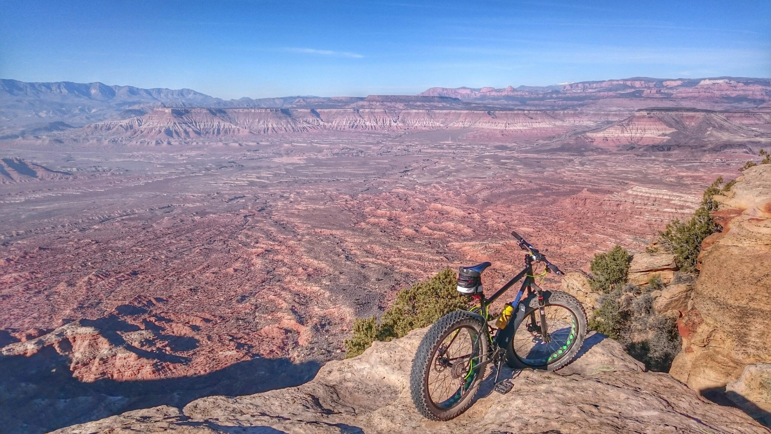 A mountain bike resting on a rocky ledge with a vast desert landscape in the background, featuring layered red and brown terrain under a clear blue sky. The scene captures the expansive beauty of the outdoors, highlighting the ruggedness of the terrain and the allure of adventure. Gooseberry Mesa mountain bike trail.