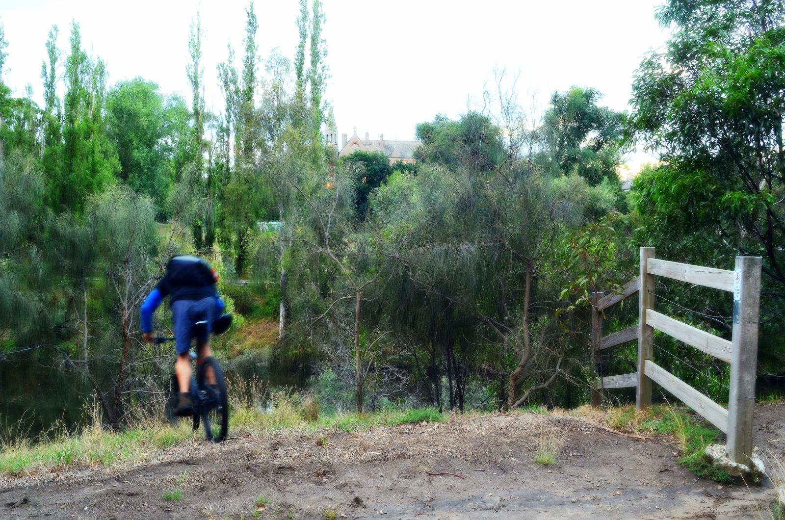 A cyclist in blue attire rides a mountain bike along a dirt path near a river, surrounded by lush greenery and trees. A wooden fence is visible on the right, and a building can be seen in the background. Yarra Trails mountain bike trail.