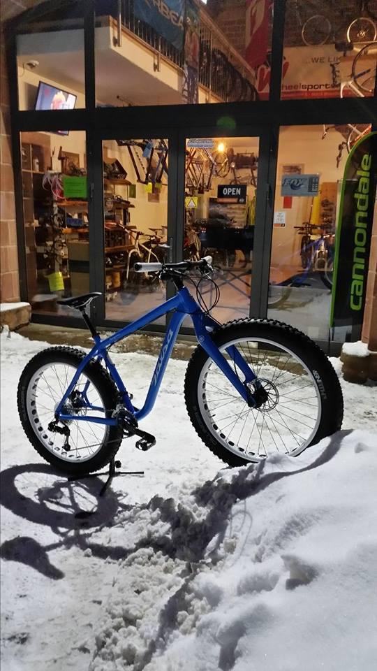 A blue mountain bike leans against a snowbank outside a bike shop with an open sign. The shop displays various bicycles through large glass windows, illuminated at night. Snow covers the ground, highlighting the winter setting.