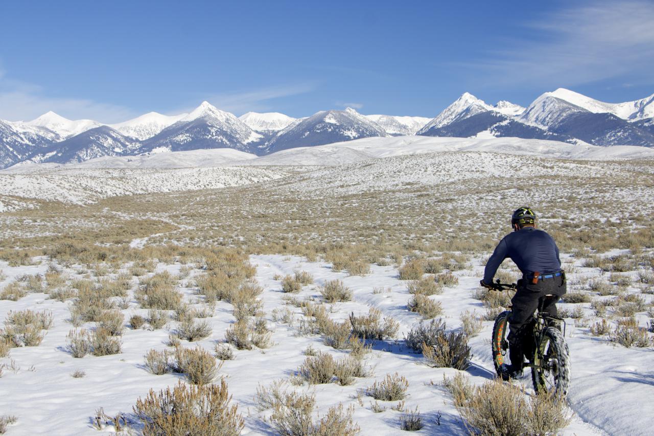 A person riding a fat bike on a snowy landscape, surrounded by sparse vegetation and snow-covered ground, with majestic snow-capped mountains in the background under a clear blue sky. Discovery Hill Trails mountain bike trail.