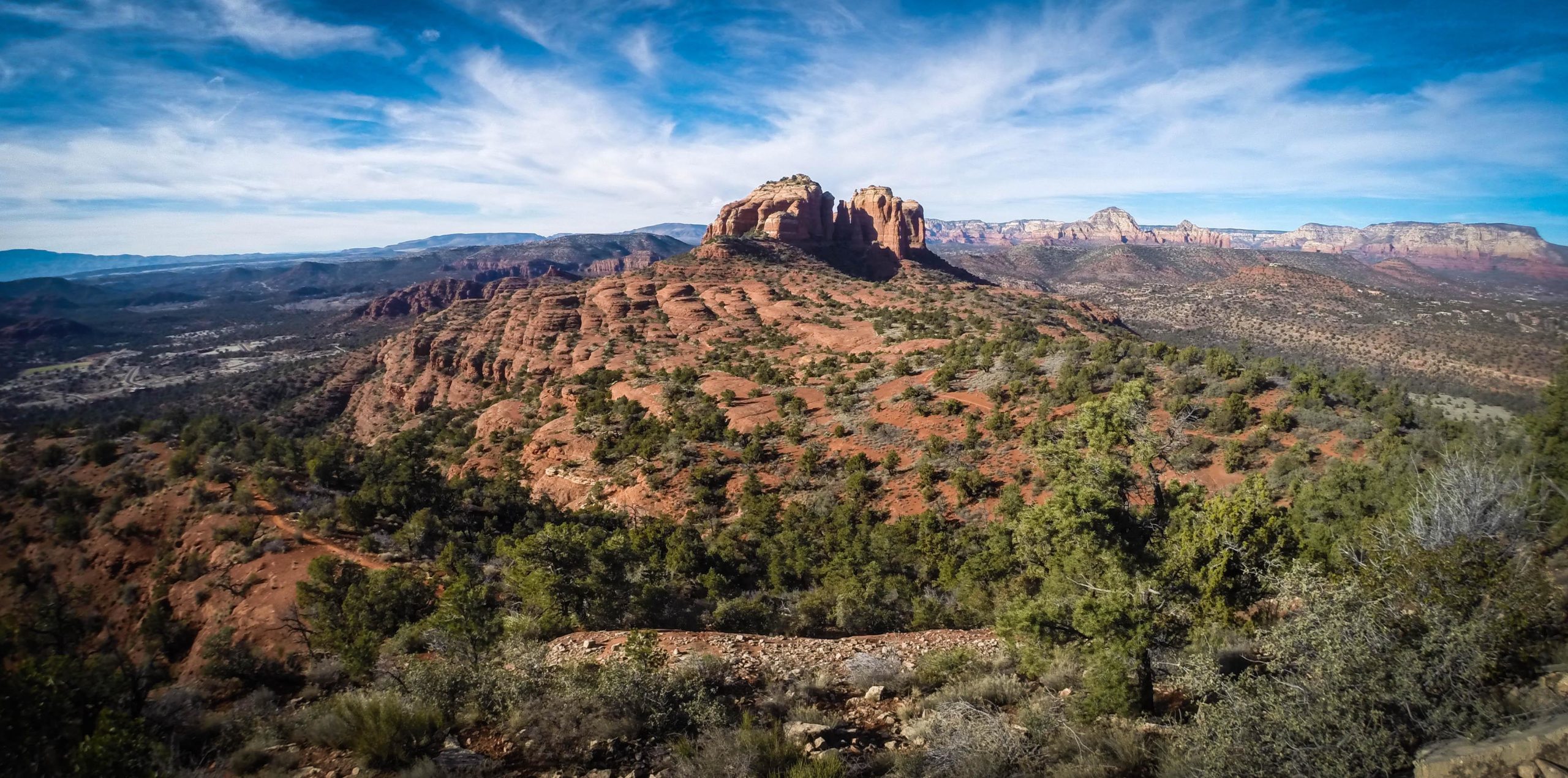 Landscape view of red rock formations and plateau with scattered green vegetation under a clear blue sky and wispy clouds. Hiline mountain bike trail.