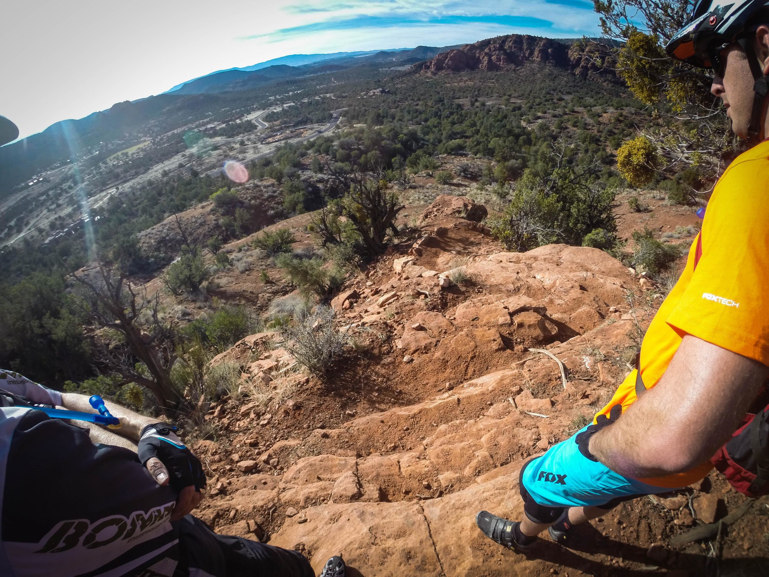 Two mountain bikers standing at the edge of a rocky hillside, overlooking a valley filled with trees and distant mountains. One biker wears a black and white jersey, while the other is dressed in an orange shirt and blue shorts. The terrain appears steep and rugged, with a clear sky above. Hiline mountain bike trail.