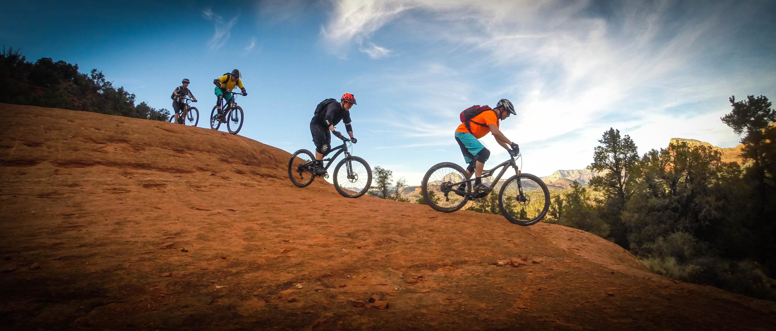 A group of four mountain bikers riding downhill on a rocky trail, surrounded by trees and blue sky with clouds. The terrain features red rock, and the bikers are wearing helmets and colorful gear, showcasing an outdoor adventure scene. Hiline mountain bike trail.