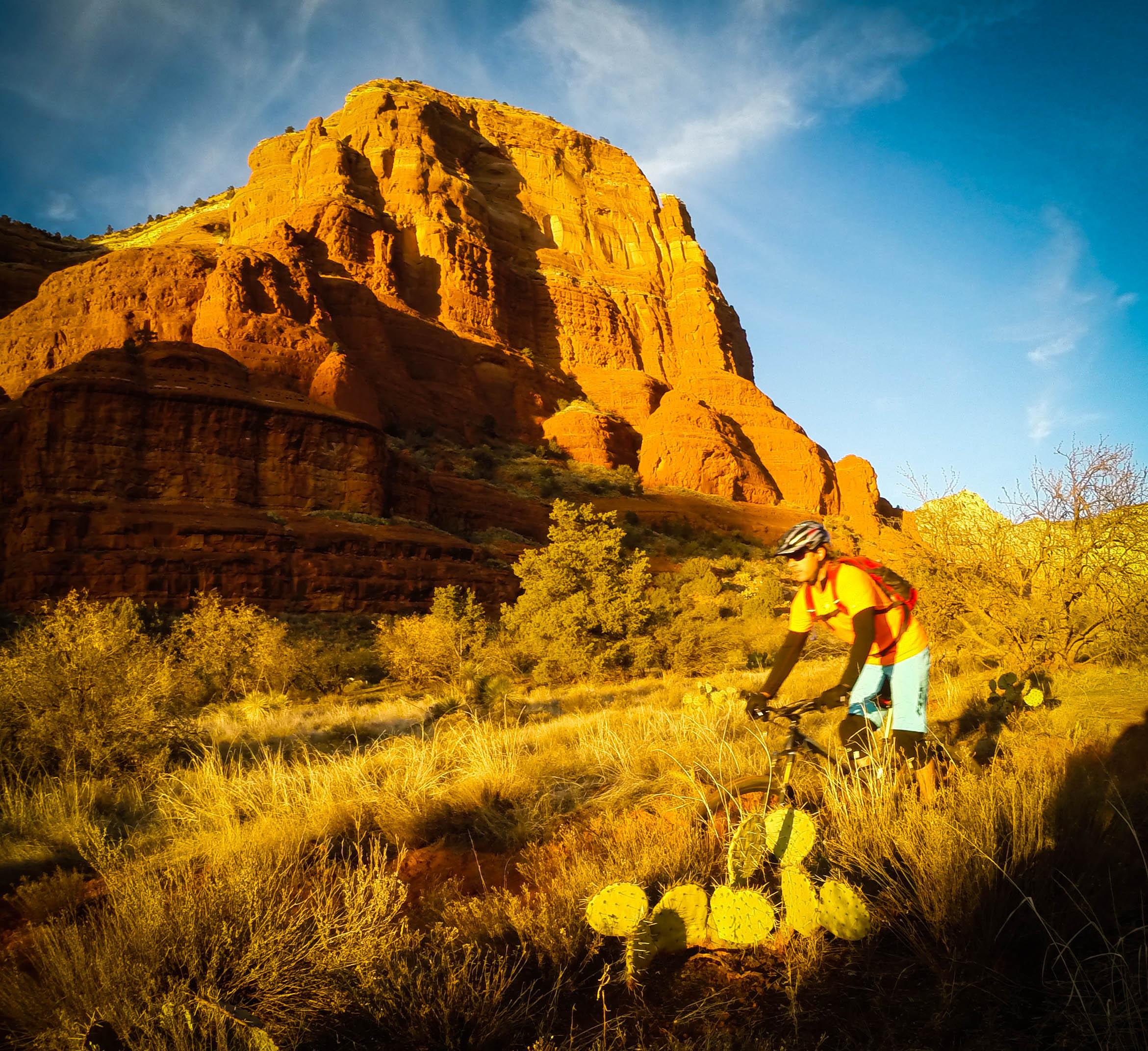 A mountain biker in bright clothing is adjusting their bike in a desert landscape, surrounded by tall red rock formations and cacti. The sun casts a warm, golden light across the scene, highlighting the vegetation and dramatic cliffs in the background. Bell Rock Trailway mountain bike trail.