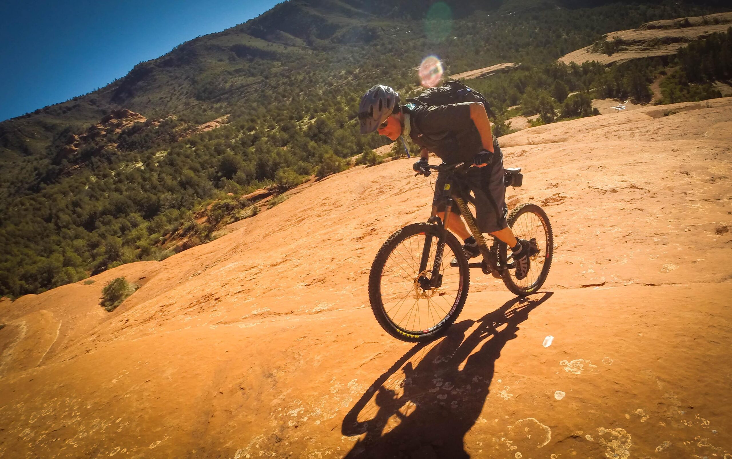 A mountain biker navigating rocky terrain on a bright day, surrounded by green hills and a clear blue sky. The biker is leaning forward on the bike as they ride down a sloped, reddish-brown surface, showcasing the adrenaline of the sport. Hangover mountain bike trail.